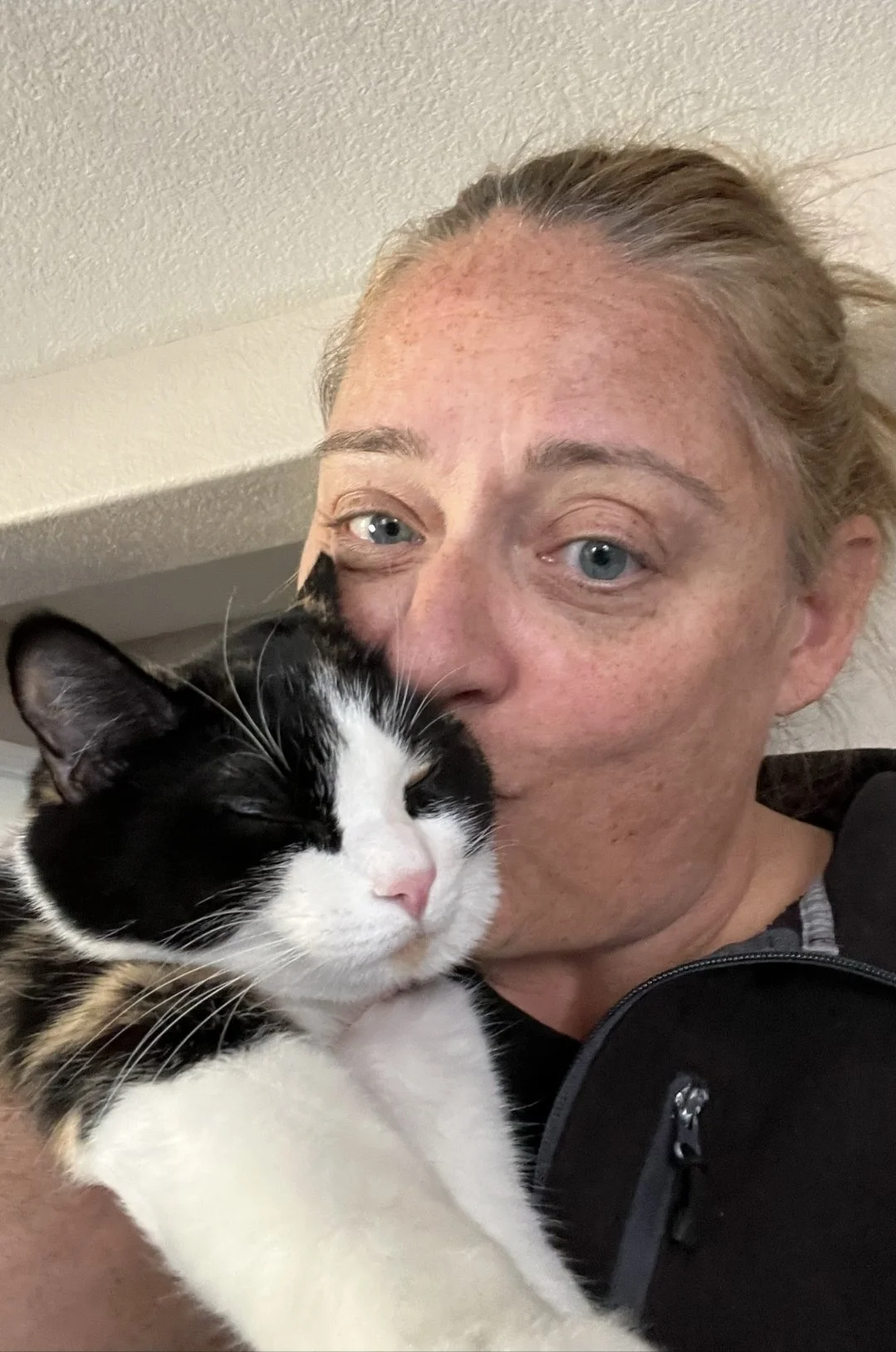 A woman with fair skin and blue eyes holding a black and white cat close to her face indoors, with a beige textured wall in the background.