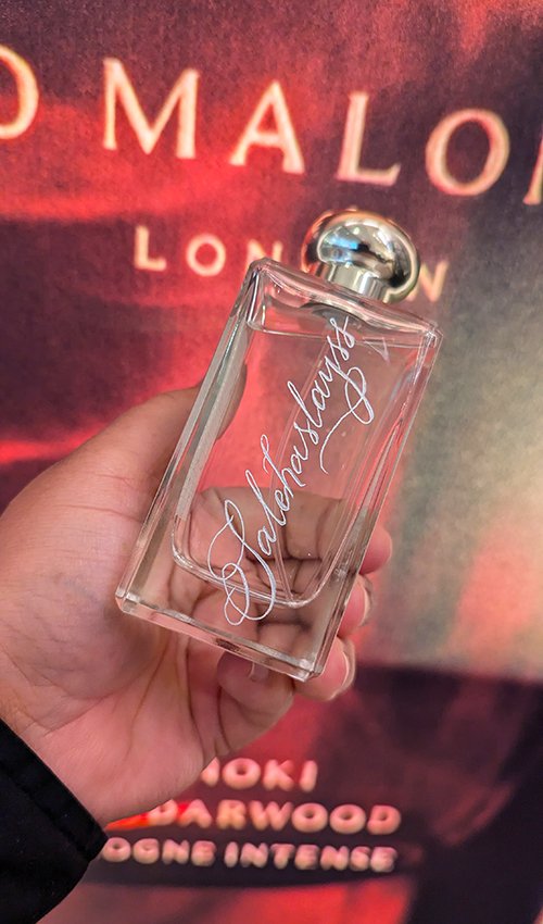A hand holding a clear perfume bottle with a silver cap, engraved with the word 'Valentino' cursive, in front of a red and black background with the words 'MALONE LONDON' and other partially visible text.