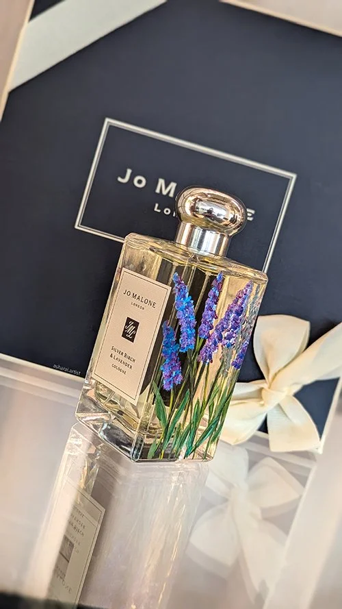 A bottle of Jo Malone Lime Basil & Mandarin cologne with painted lavender flowers on it, placed in front of a Jo Malone box with a cream-colored ribbon.