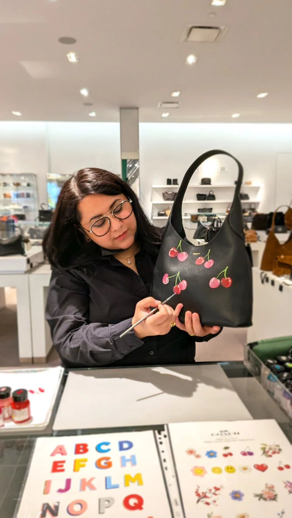 A woman painting cherry illustrations on a black handbag in a retail store, with colorful alphabet and floral designs on display in front of her.