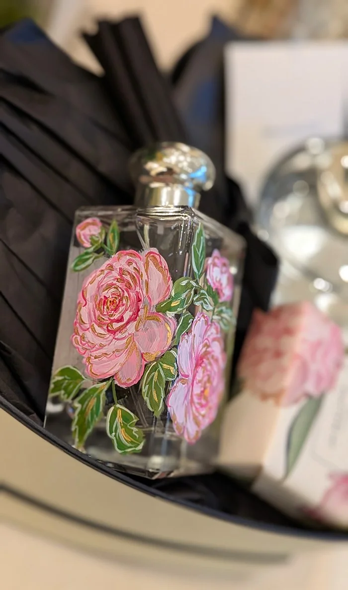 Close-up of a glass perfume bottle with pink rose flower design, black ribbon, and some blurred objects in the background.