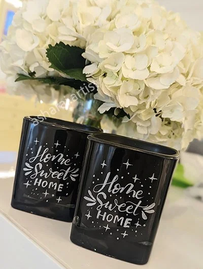 Two black mugs with white writing that says "Home Sweet Home" and decorative stars and leaves, placed in front of a bouquet of white hydrangeas.