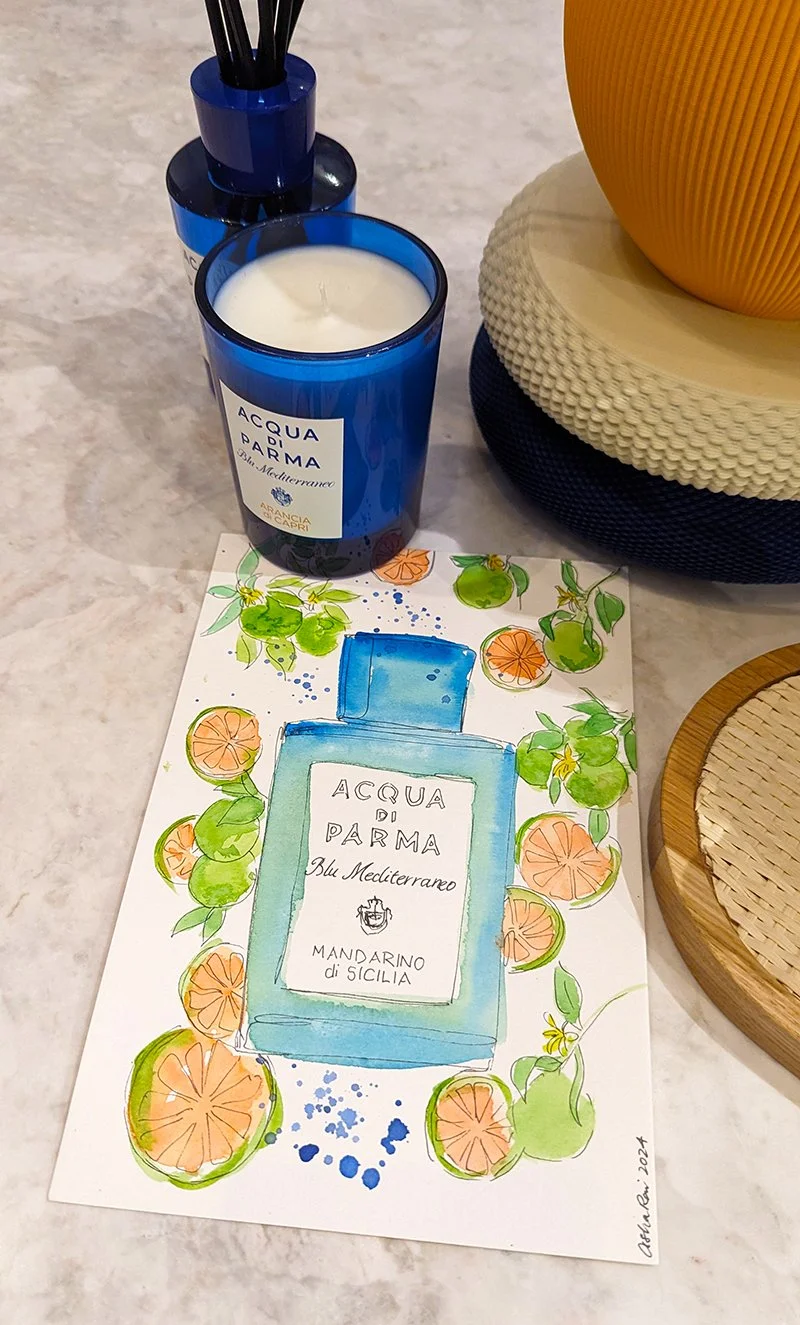 A blue candle labeled "Acqua di Parma Blu Mediterraneo Mandarino di Sicilia" placed on a table next to a colorful illustration of the perfume bottle and mandarin oranges, with decorative bowls nearby.