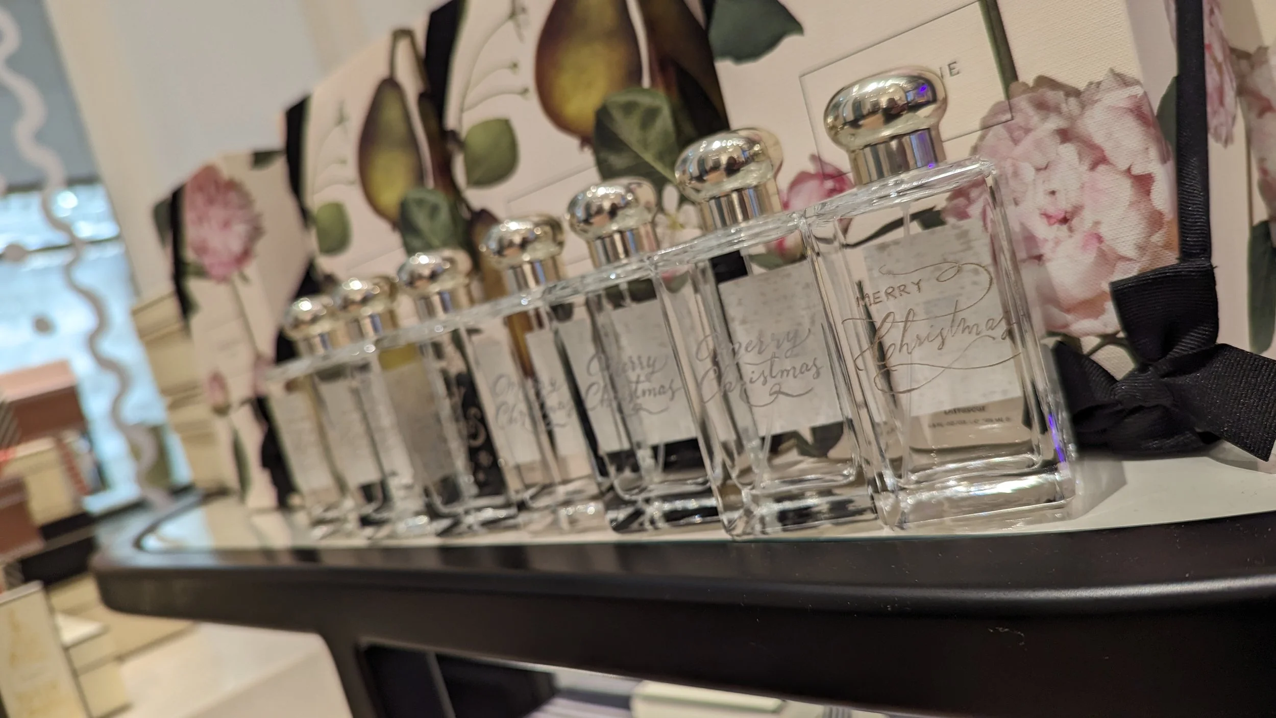 Set of clear glass perfume bottles with silver caps labeled "Merry Christmas" arranged on a reflective surface, with a floral and fruit decorative box in the background.