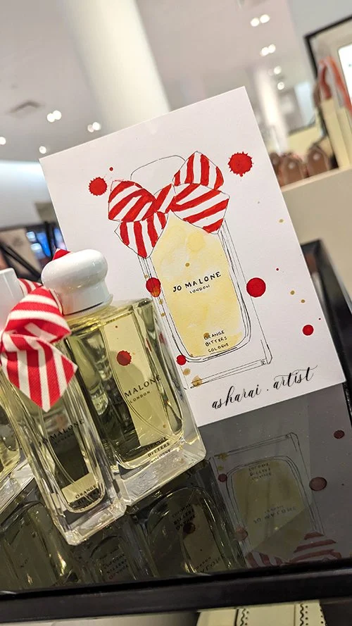 Live watercolor illustration by AshaRai of a Jo Malone limited edition Orange Bitters perfume bottle decorated with red-and-white striped ribbon bows, created at the Jo Malone counter at Yorkdale.