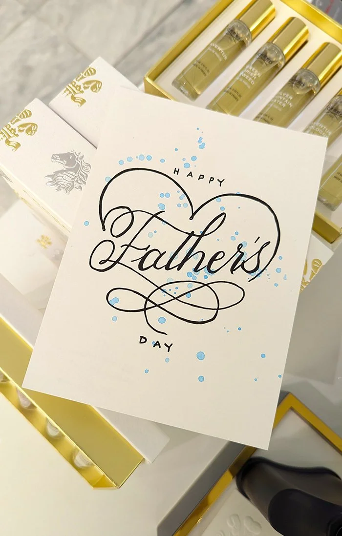 A greeting card with the text 'Happy Father's Day' written in elegant black cursive, decorated with blue ink splashes, placed on a display with small perfume bottles and boxes.