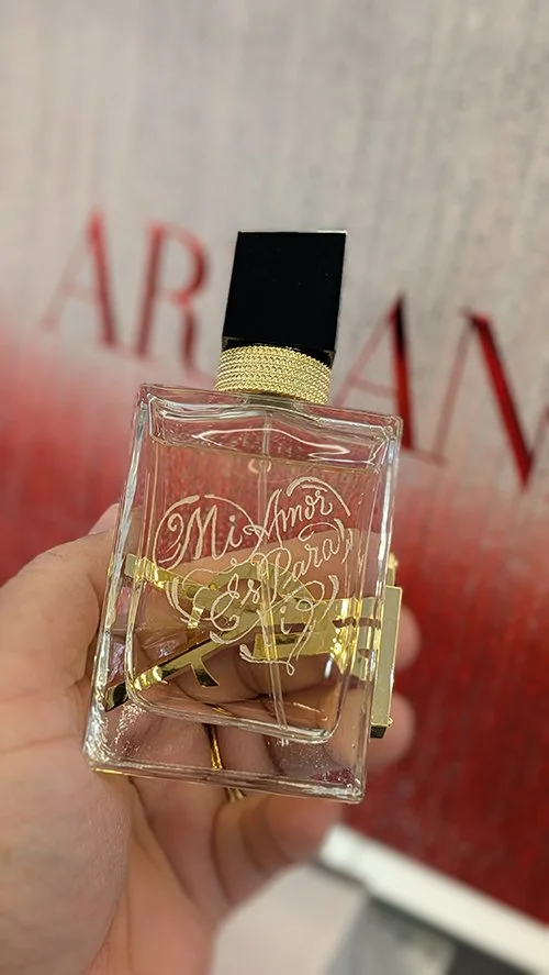 A hand holding a bottle of perfume with a black cap and gold accents, displaying the words 'Mama' and 'Pura' etched on the front.
