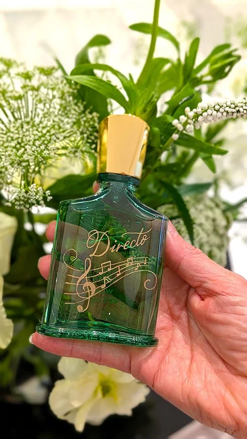 A hand holding a green perfume bottle with a gold cap, decorated with gold musical notes and the word 'Directo', surrounded by white flowers and green leaves.