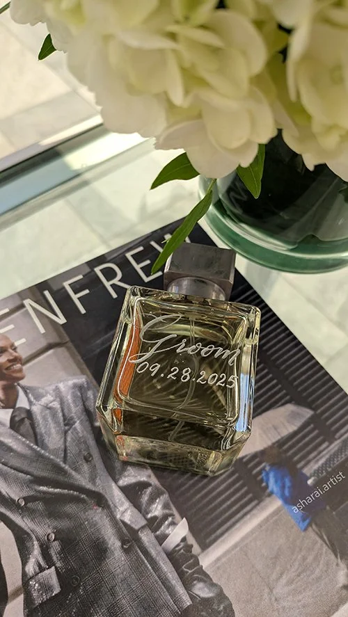 A glass perfume bottle labeled 'Groom 09.28.2023' in cursive, placed on a magazine with a man in a gray suit, next to a vase of white hydrangeas on a glass surface.