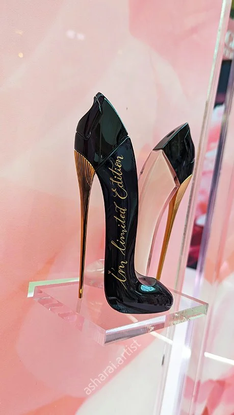 A pair of high-heeled shoes on a clear display shelf, with a pink background. The shoes are black with gold accents, shaped like elegant stilettos, and one shoe has the words "Uninhibited Admission" written on it.
