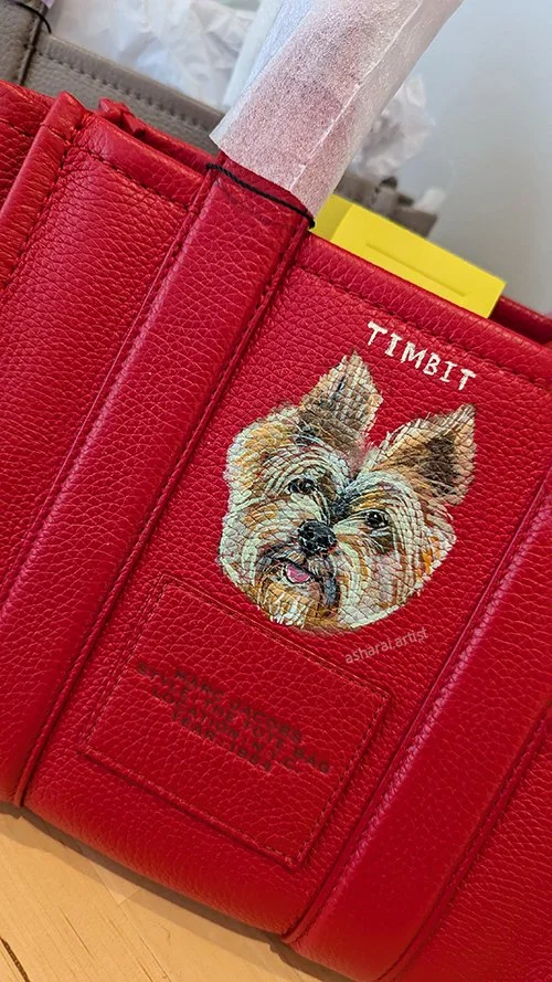 Close-up of a red textured Marc Jacobs tote bag with a customized pet portrait of a small dog, labeled "Timbit" in Yorkdale mall at Marc Jacobs Boutique by events artist Asha Rai