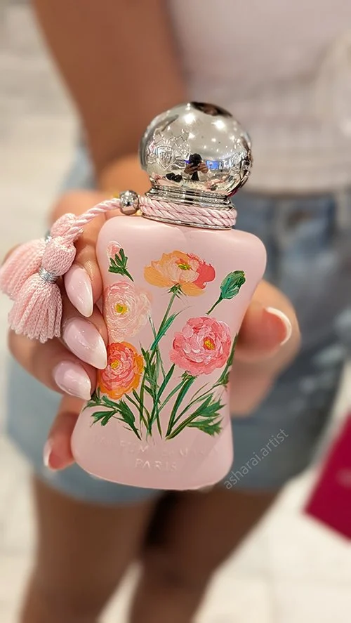 A woman holding a pink perfume bottle with floral artwork and a silver cap, with a pink tassel attached, against a blurred background.