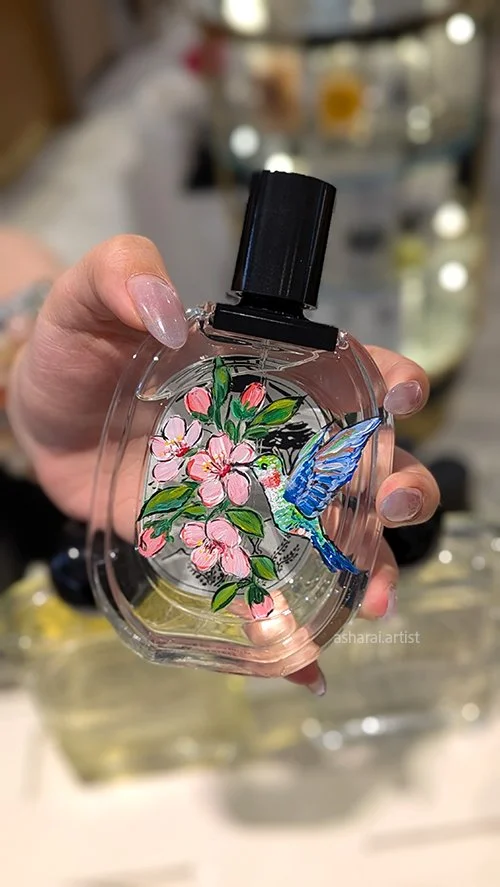 Diptyque perfume bottle featuring hand-painted pink flowers and a blue hummingbird, showcasing live bottle painting at Holt Renfrew Yorkdale.