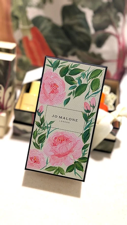 Jo Malone London display card with pink floral design and green leaves, standing on a white surface in a store.