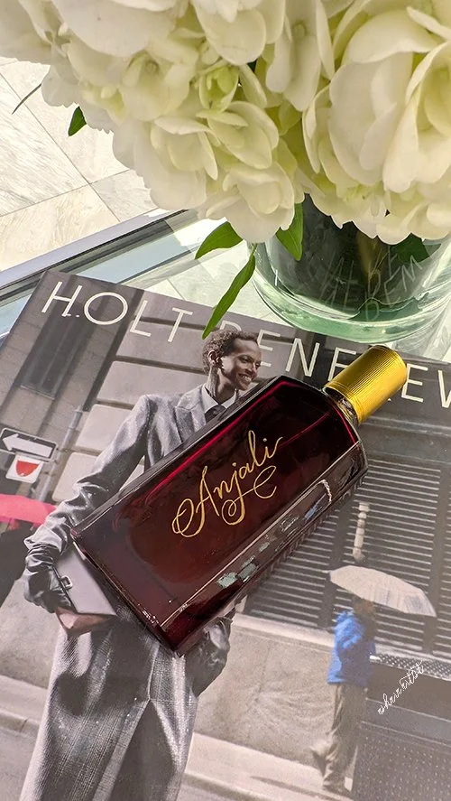 A magazine featuring a woman in a gray suit with a book under one arm, a bottle with a gold cap labeled 'Anjali,' and a vase of white hydrangea flowers on top.