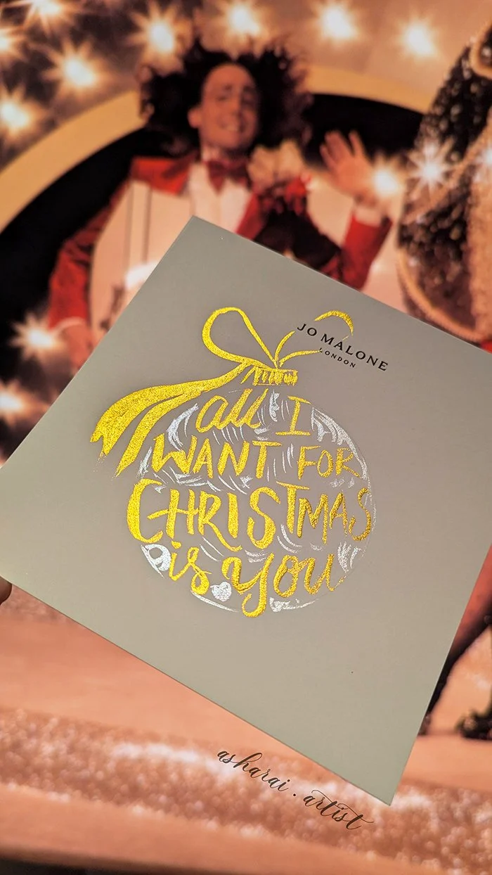 A Christmas card with a gold-embossed illustration of a Christmas ornament and the message 'All I want for Christmas is you.' The card is from Jo Malone London and painted by Live event artist .