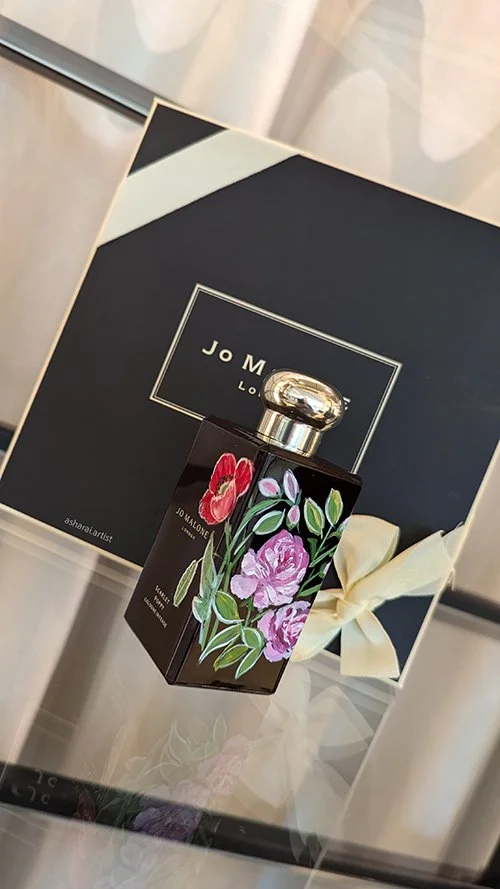A Jo Malone London perfume bottle with a floral design, placed in front of a Jo Malone London box with a cream-colored ribbon, on a reflective surface.
