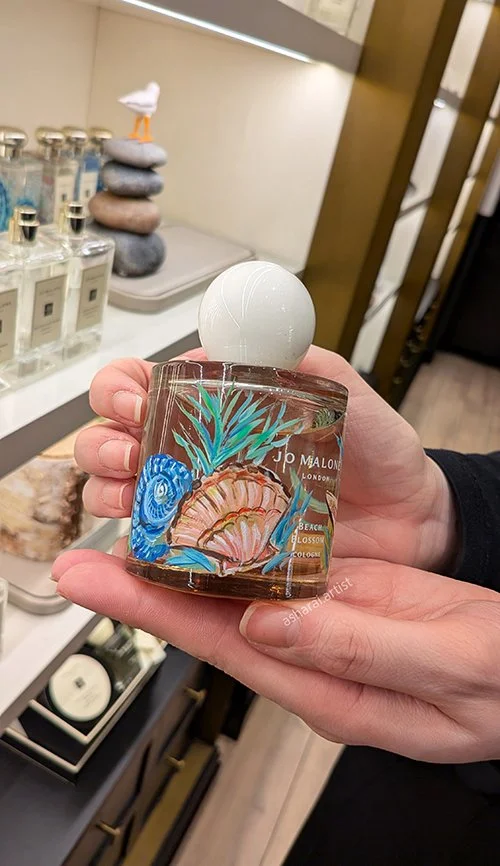 Jo Malone London Beach Blossom perfume bottle featuring a hand painted seashell and ocean motif, with a white spherical cap.Jo Malone London Yorkdale.