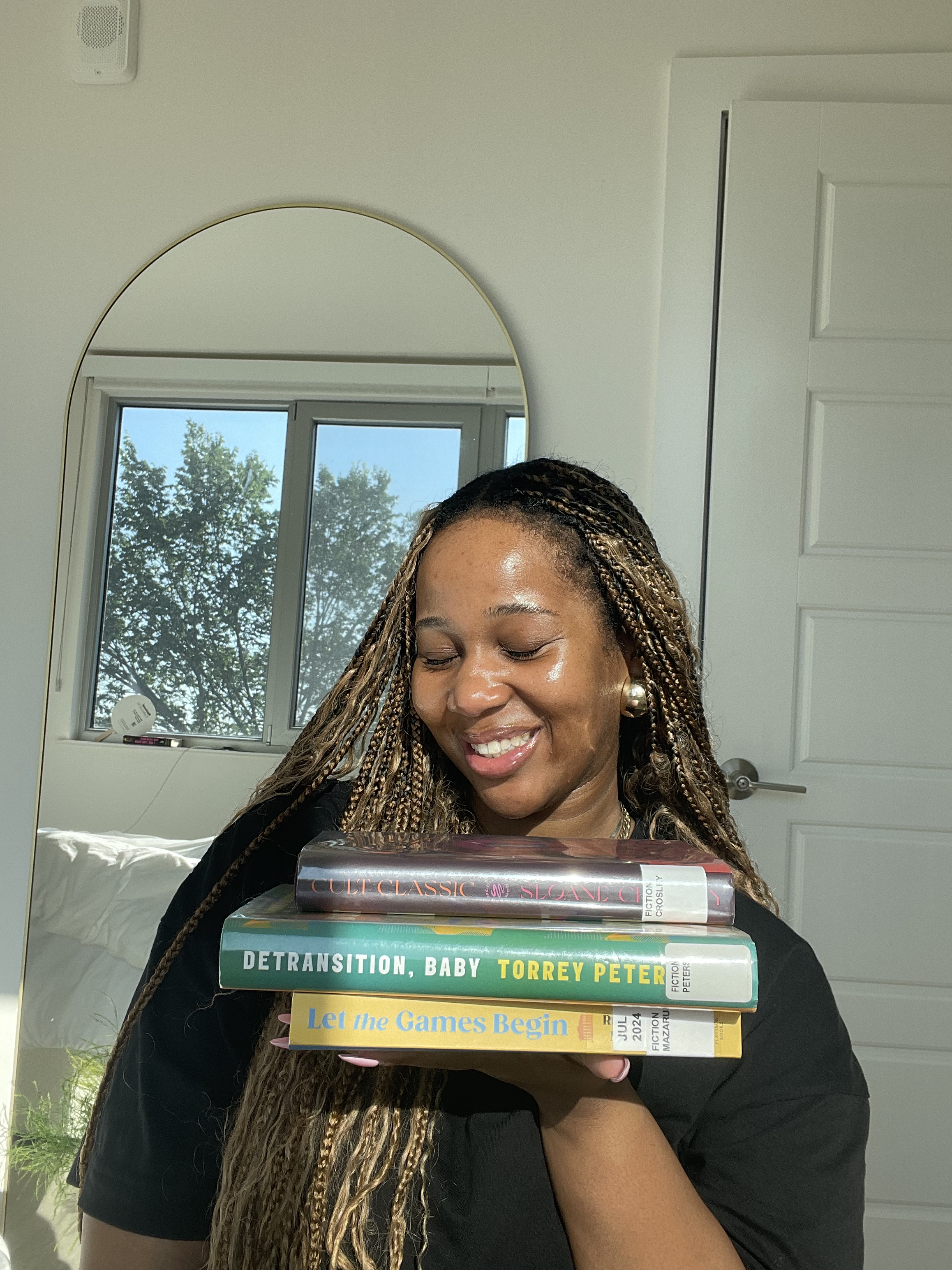 A woman with braided hair and earrings holds a stack of books in front of her face, smiling with her eyes closed, inside a room with a window showing trees outside.