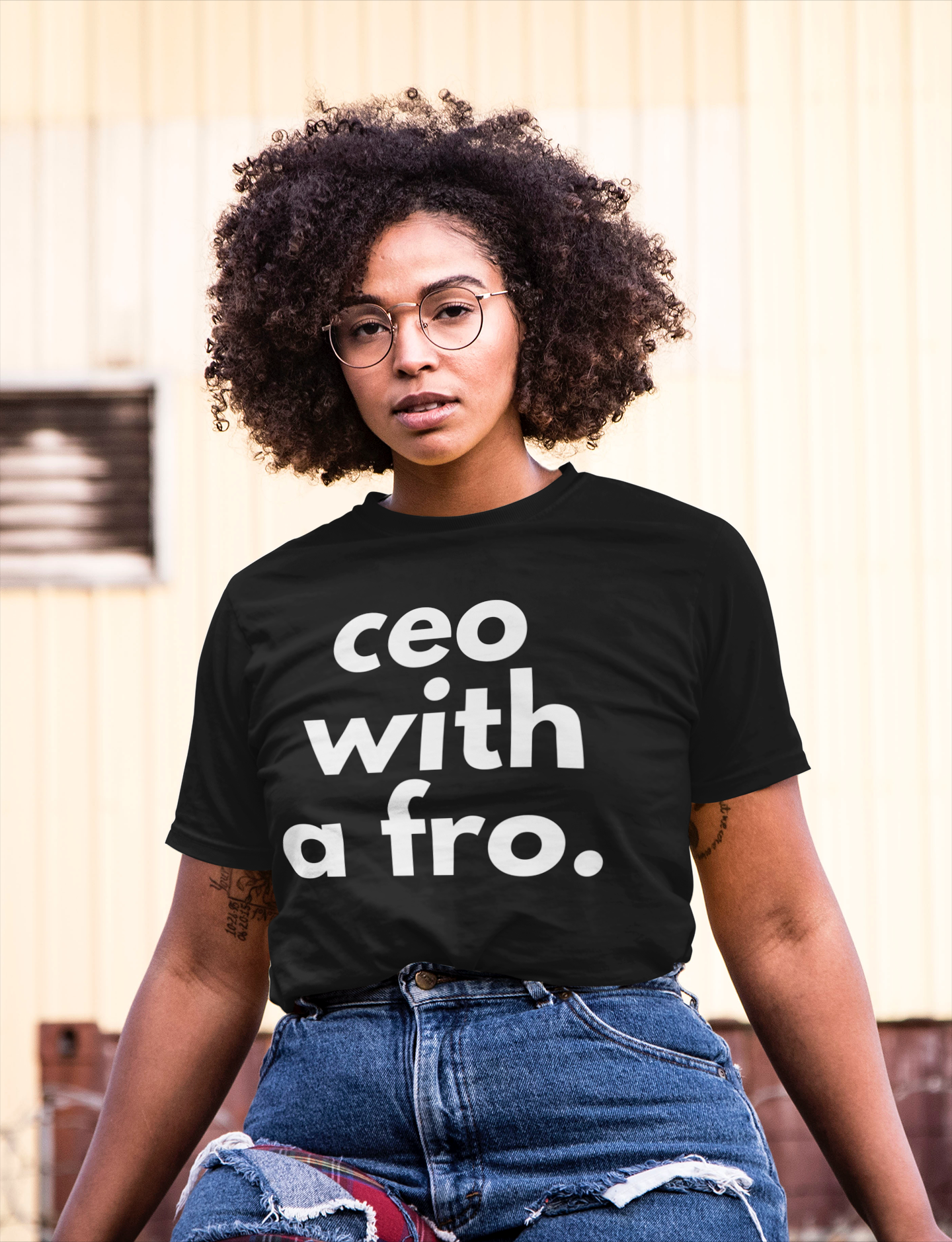 A woman with curly hair, wearing glasses and a black T-shirt with the text "CEO with a fro," seated outdoors against a background of a yellow building and a vent.