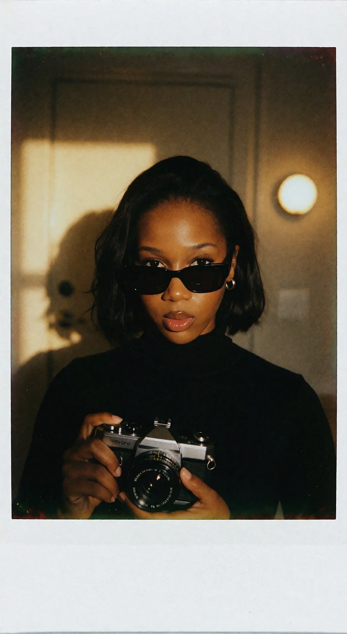 A woman with dark hair wearing sunglasses and a black turtleneck, holding a vintage camera, taking a self-portrait in a mirror.