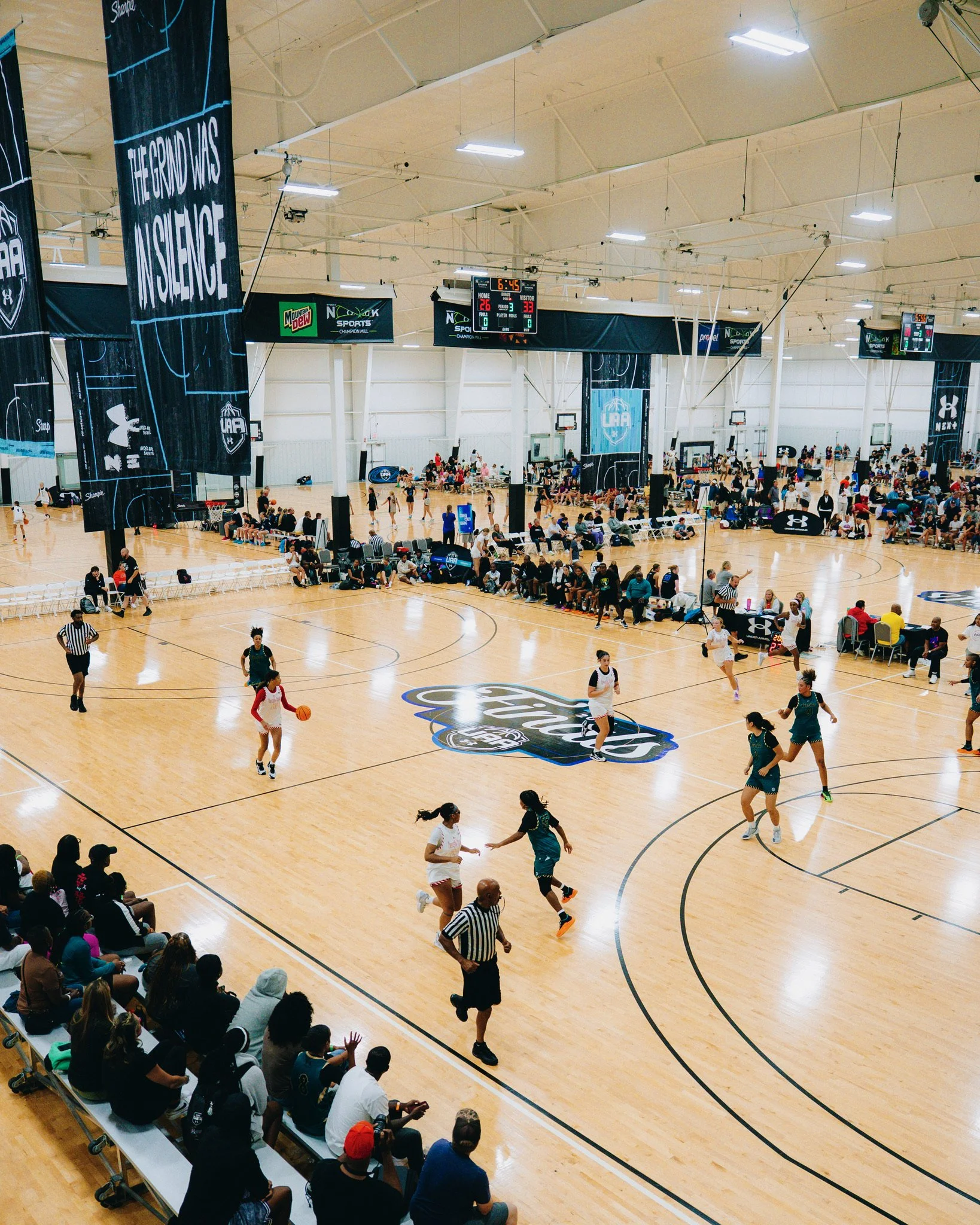 An indoor basketball game with players on the court, spectators on the sidelines, and two electronic scoreboards hanging from the ceiling, indicating the game time and scores.