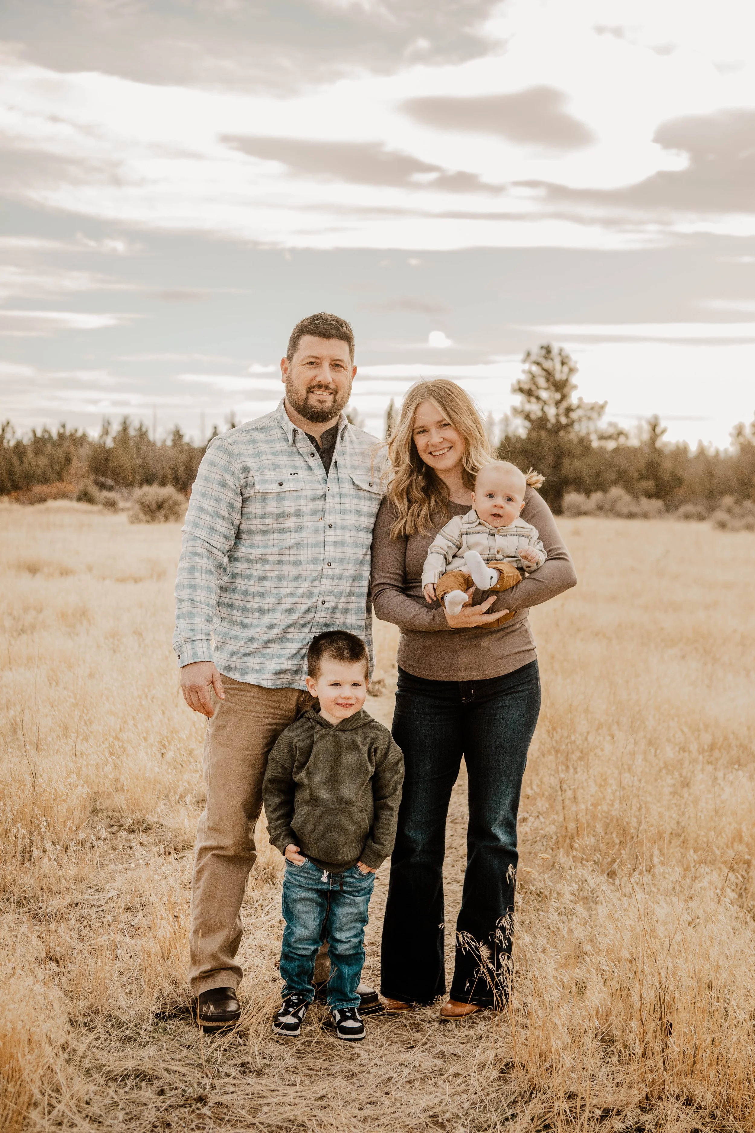 A family of four standing outdoors in a field of dry grass, with a cloudy sky and trees in the background. The mother is holding a baby, and two young boys are standing in front of them.