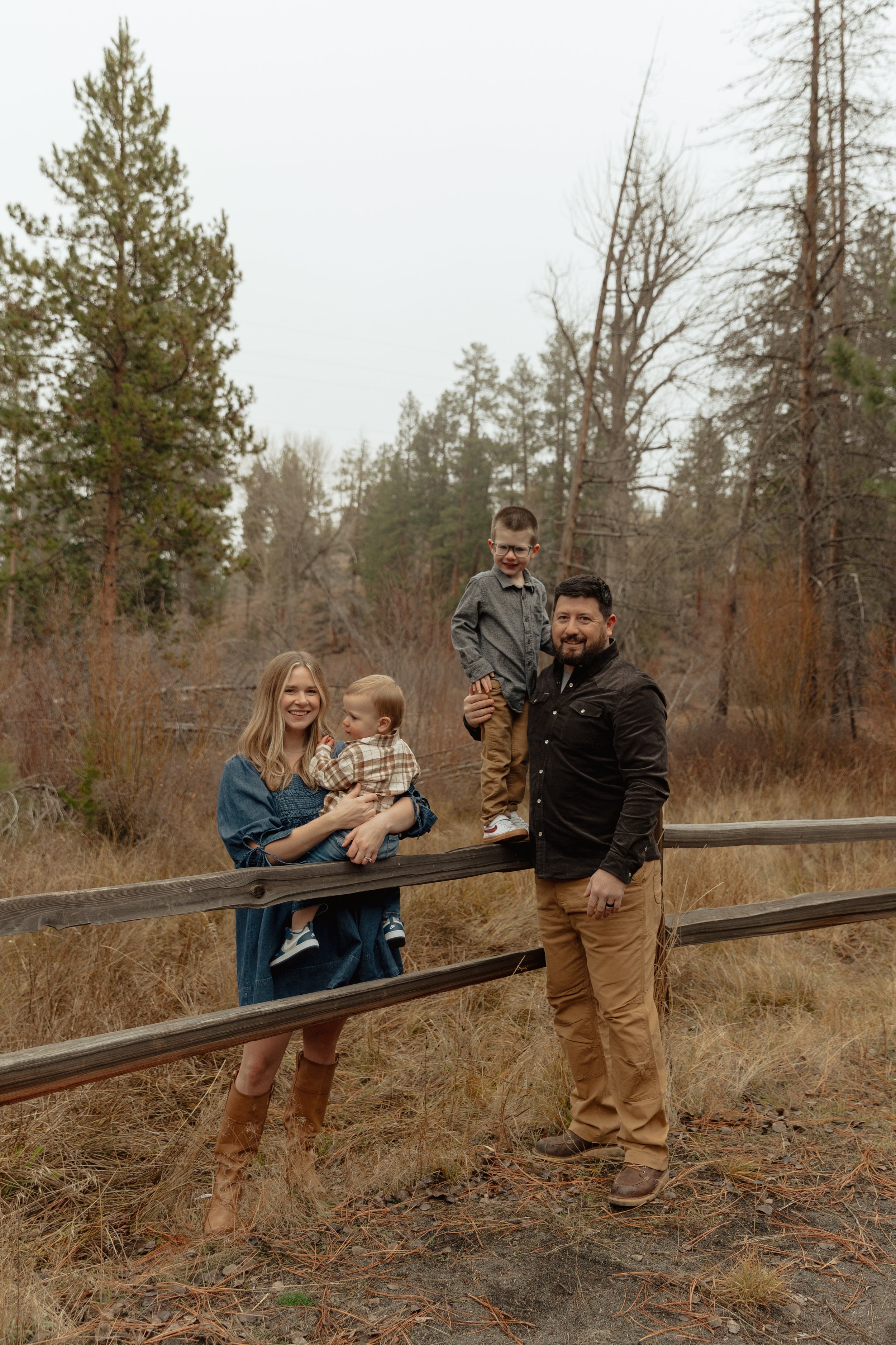 A family of five outdoors in a forest clearing during fall, with three children and two adults, standing near a wooden fence.