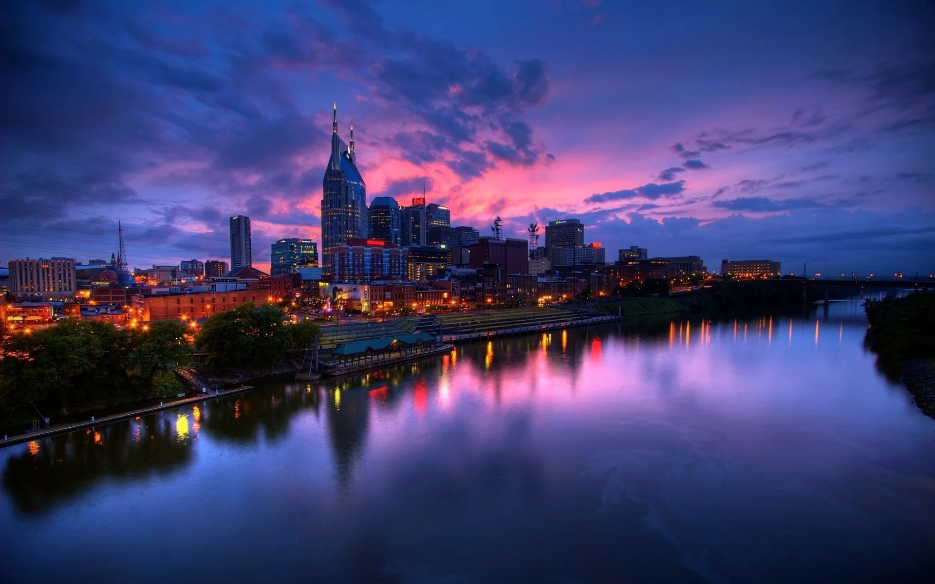 Nighttime Nashville skyline with modern skyscrapers, including the tallest one with spires, illuminated by city lights and reflections on a calm river, under a colorful sunset sky with clouds.
