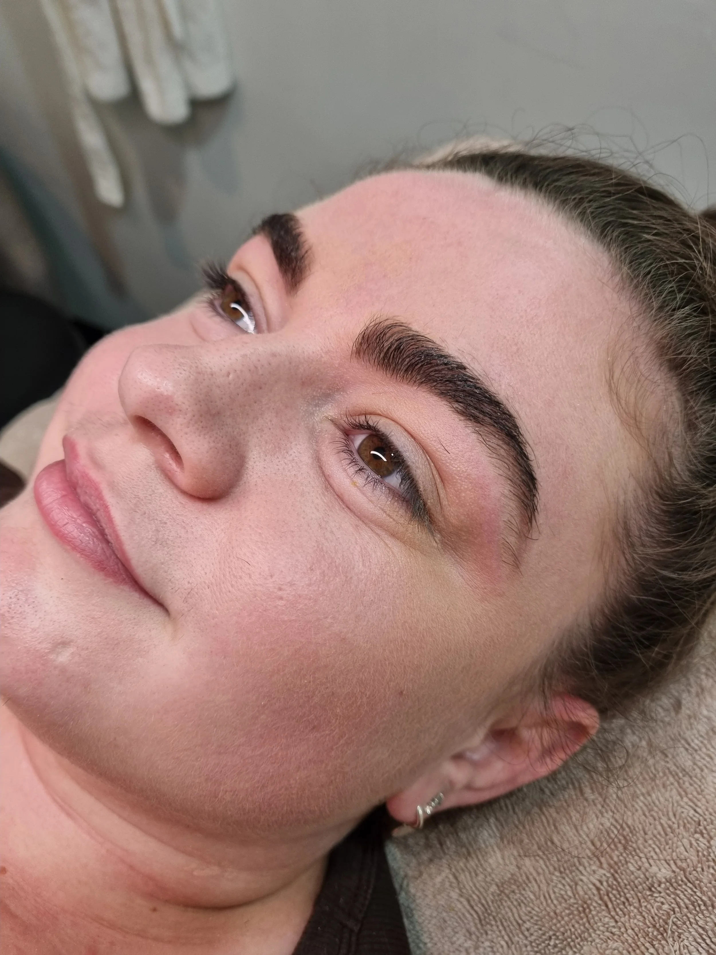 Close-up of a woman's face showing fresh, groomed eyebrows, brown eyes, and pink lips, lying on a beige towel.