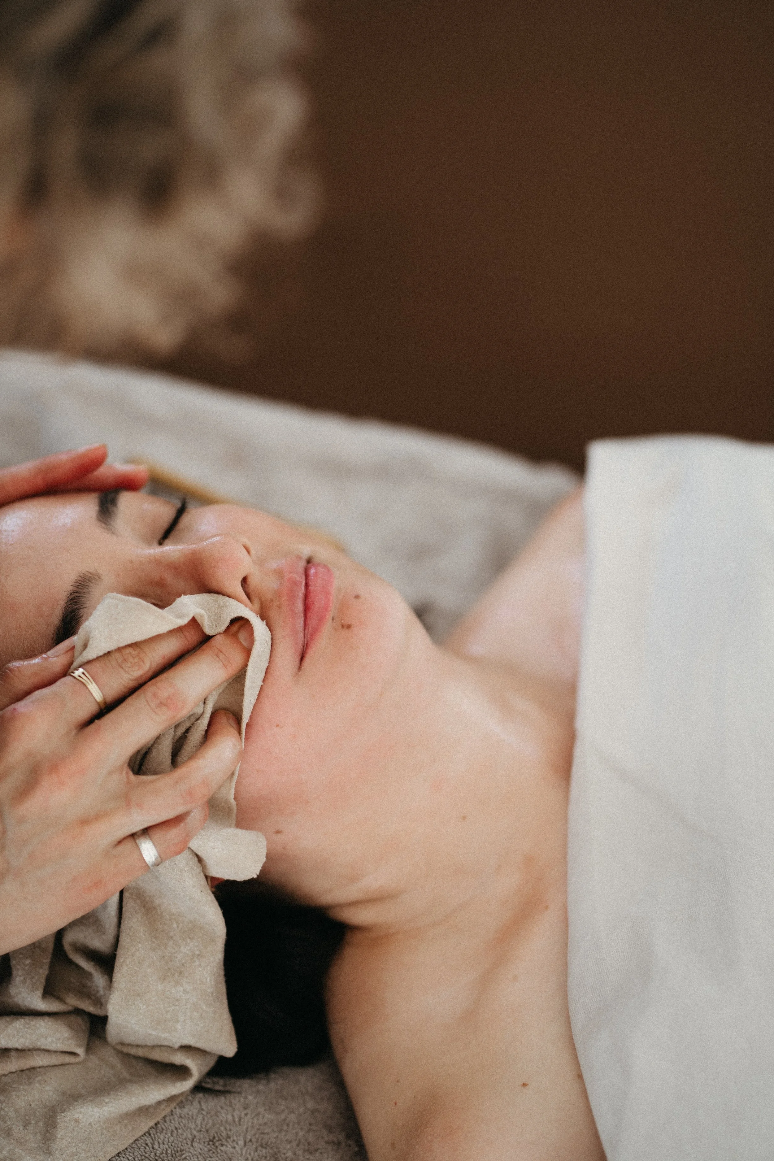 Person receiving facial massage or skincare treatment, lying down with closed eyes, person's face being gently touched by another person.