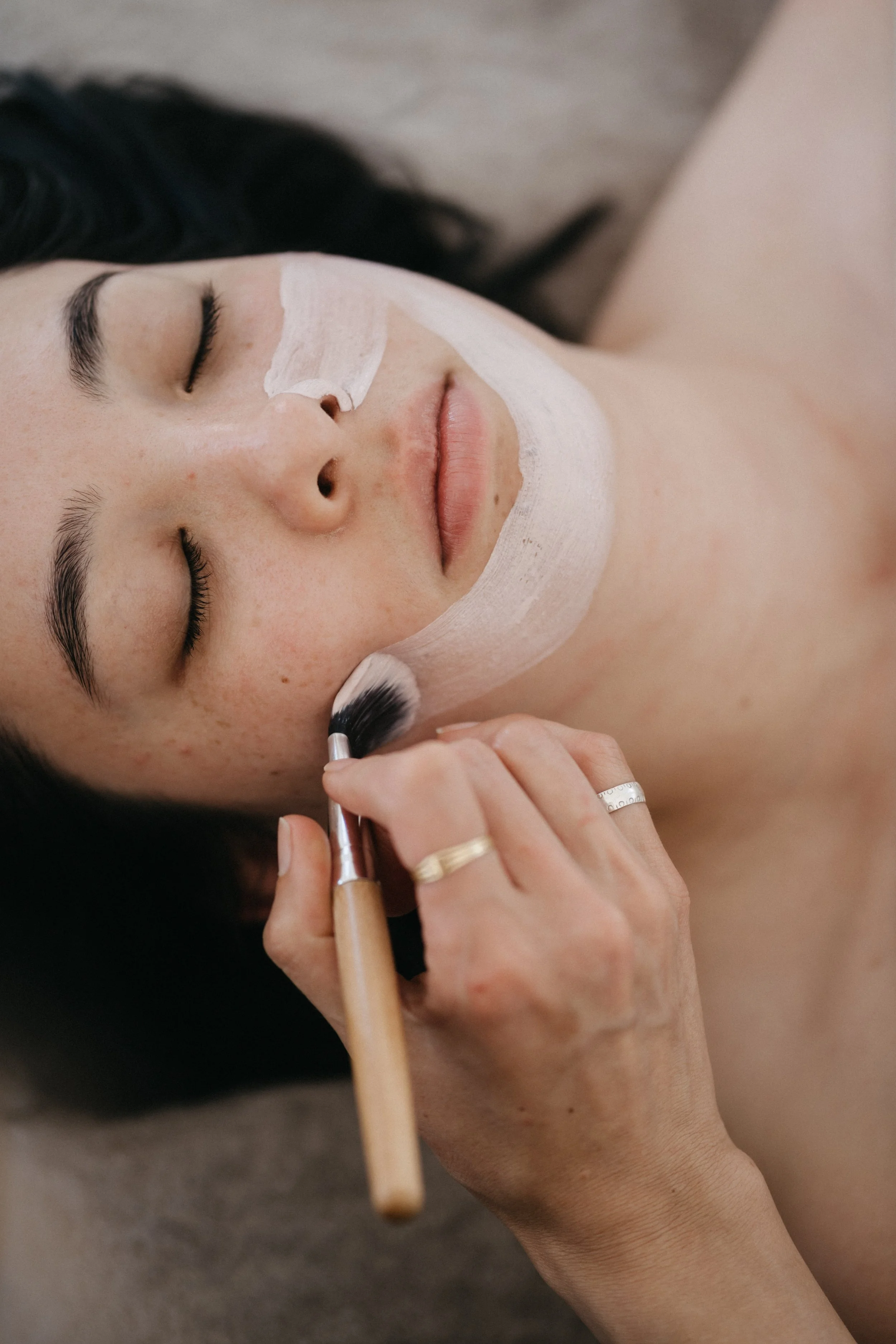 A woman with dark hair and fair skin receives a facial treatment. She has a white mask applied on her face and is lying down with closed eyes. A person is applying makeup with a brush near her cheek.