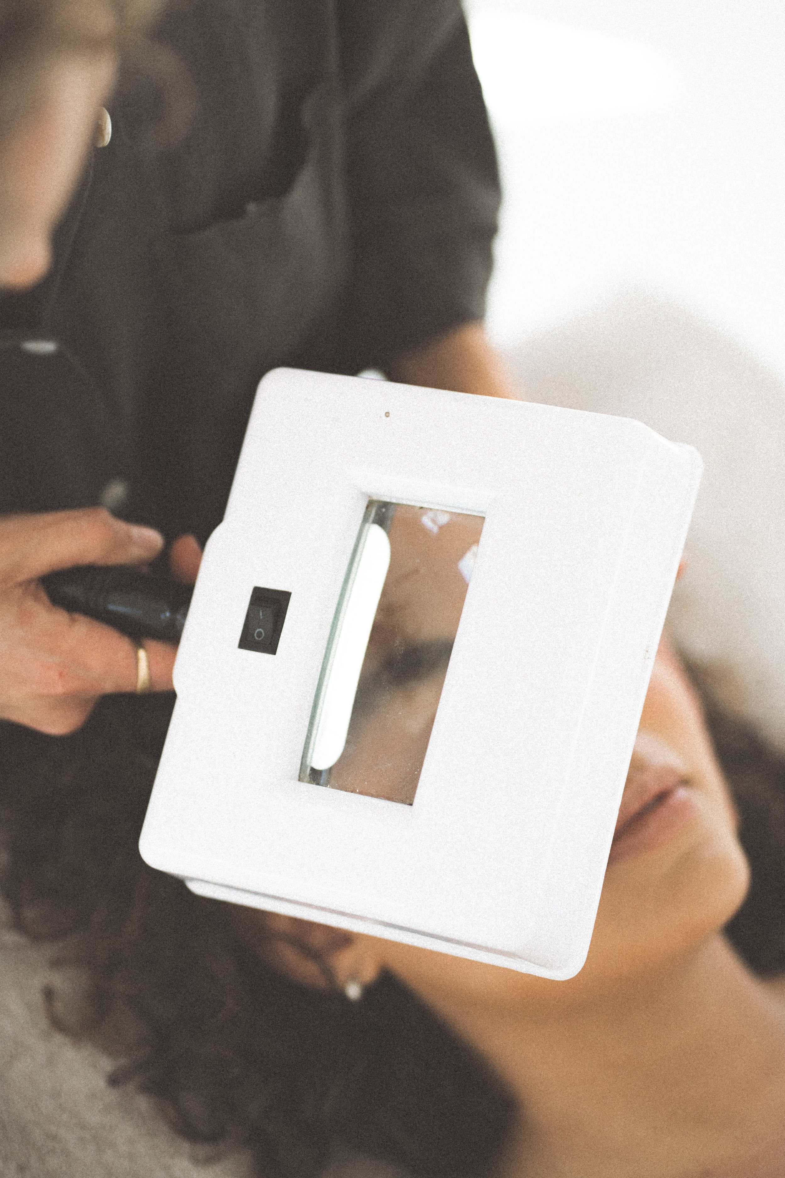 A person holding a white table mirror and taking a selfie, with the reflection showing part of their face and dark curly hair.