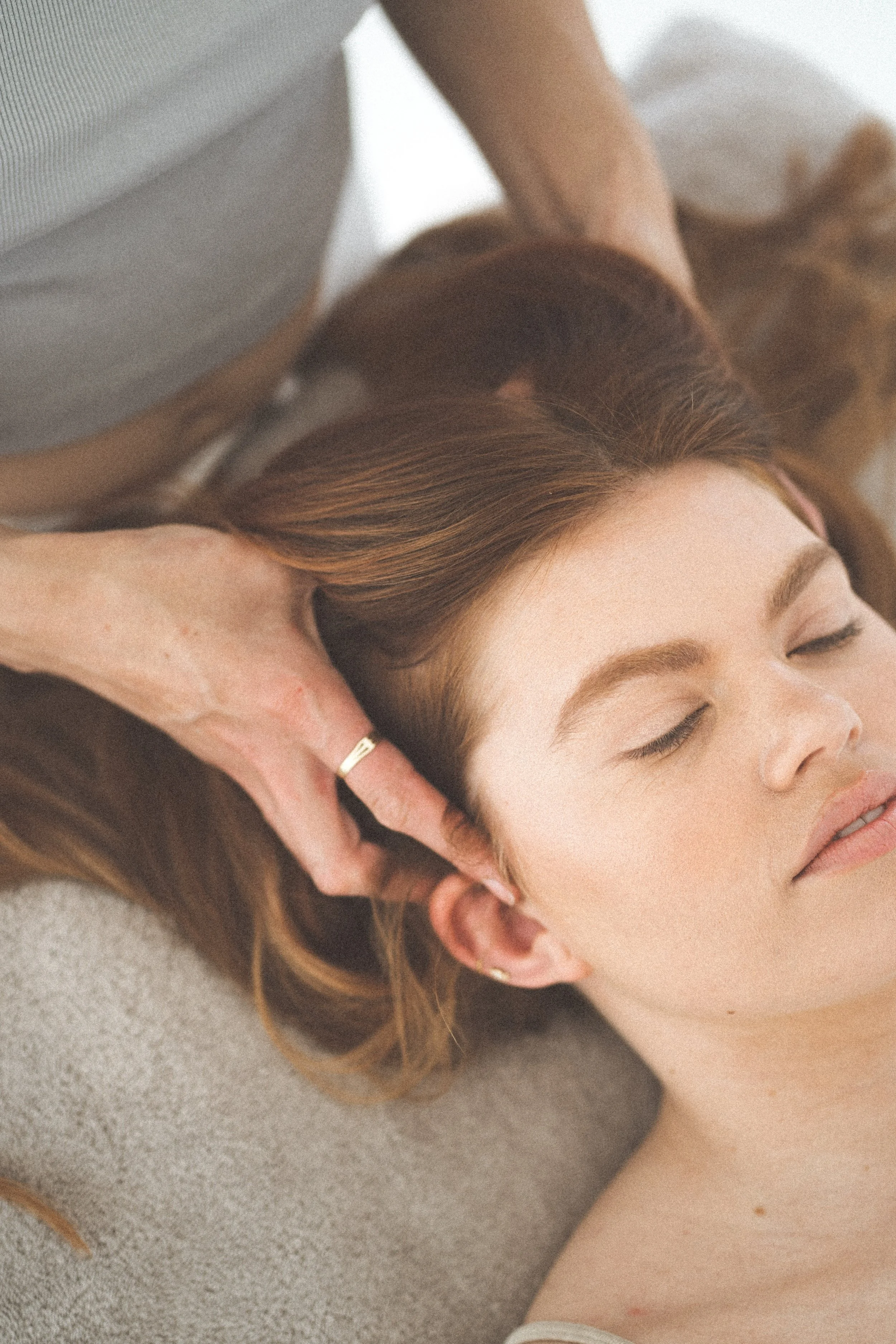 A woman with red hair is lying on her back with her eyes closed, receiving a massage or facial treatment from a person whose hand is resting on her forehead. The woman appears relaxed and calm.