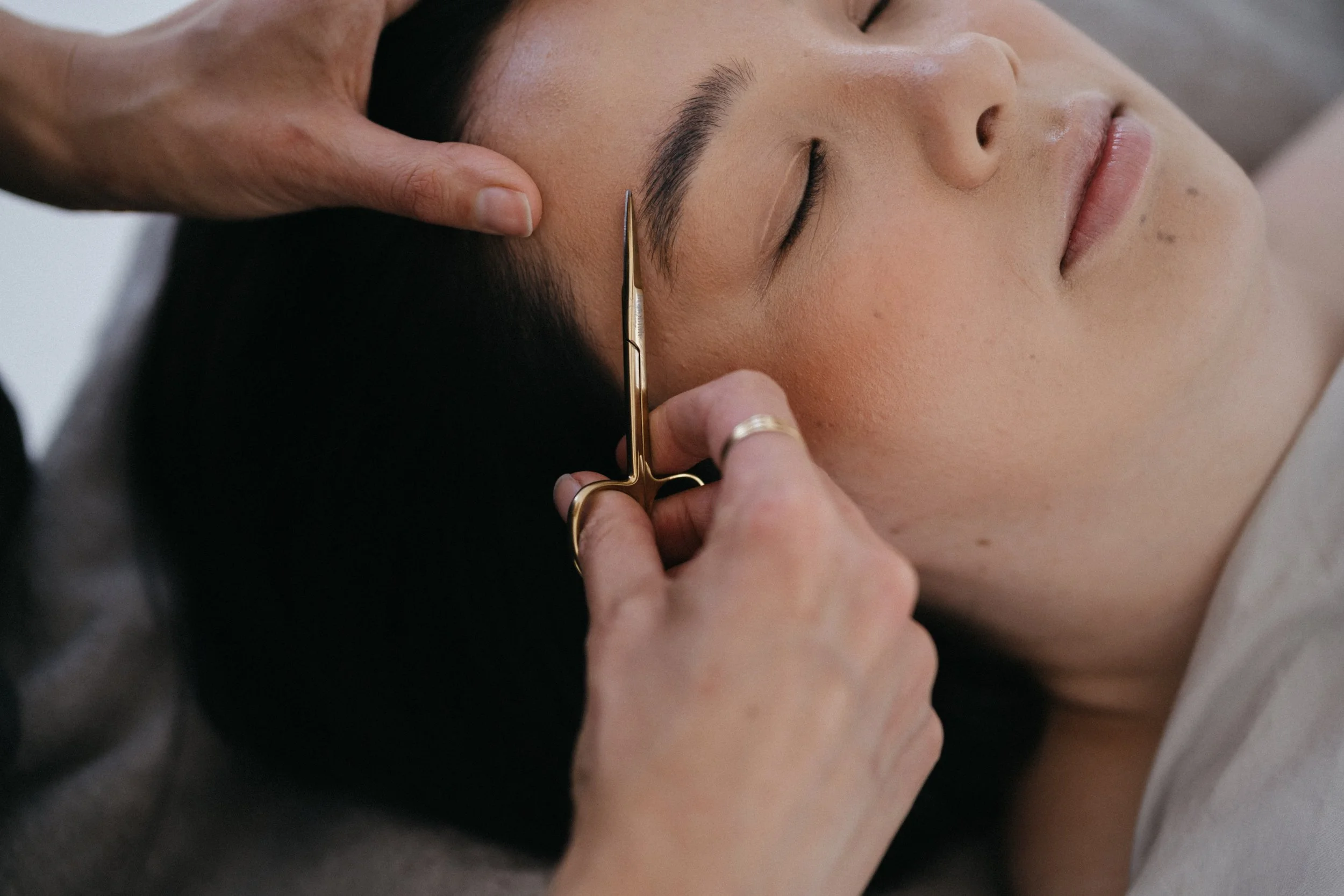 A person receiving an eyebrow grooming treatment, with a stylist using tweezers on the person's eyebrow.