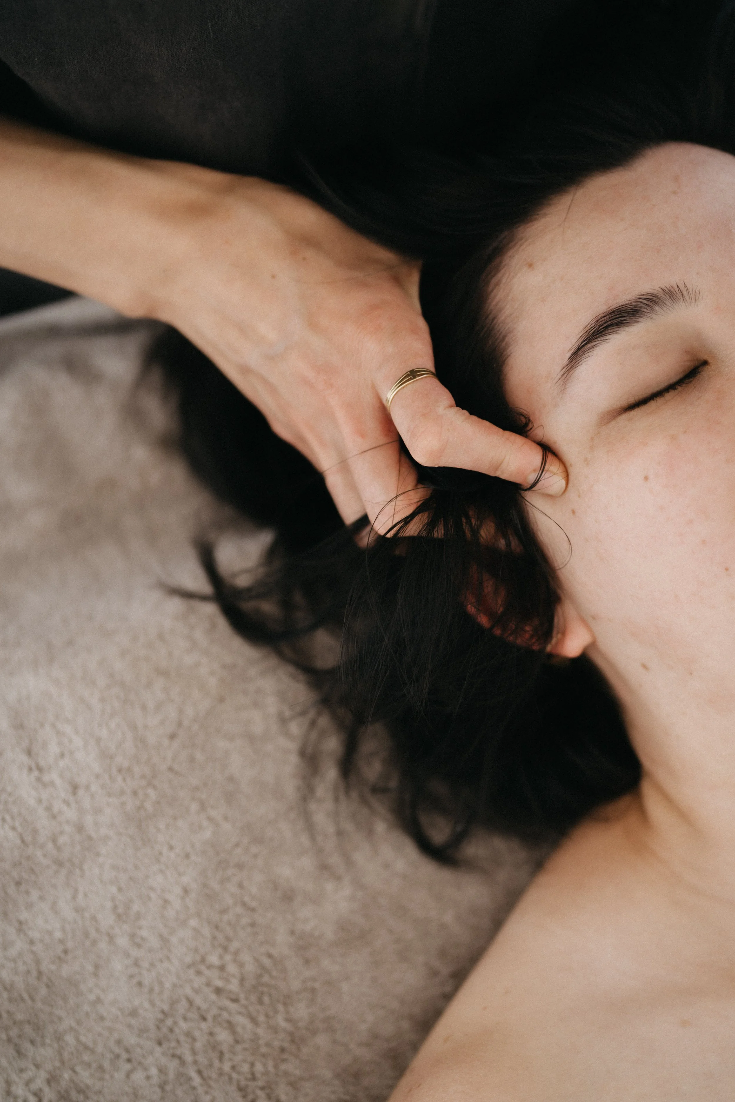A woman with closed eyes and dark hair lying on a beige carpet, with a hand near her temple, wearing a gold ring.