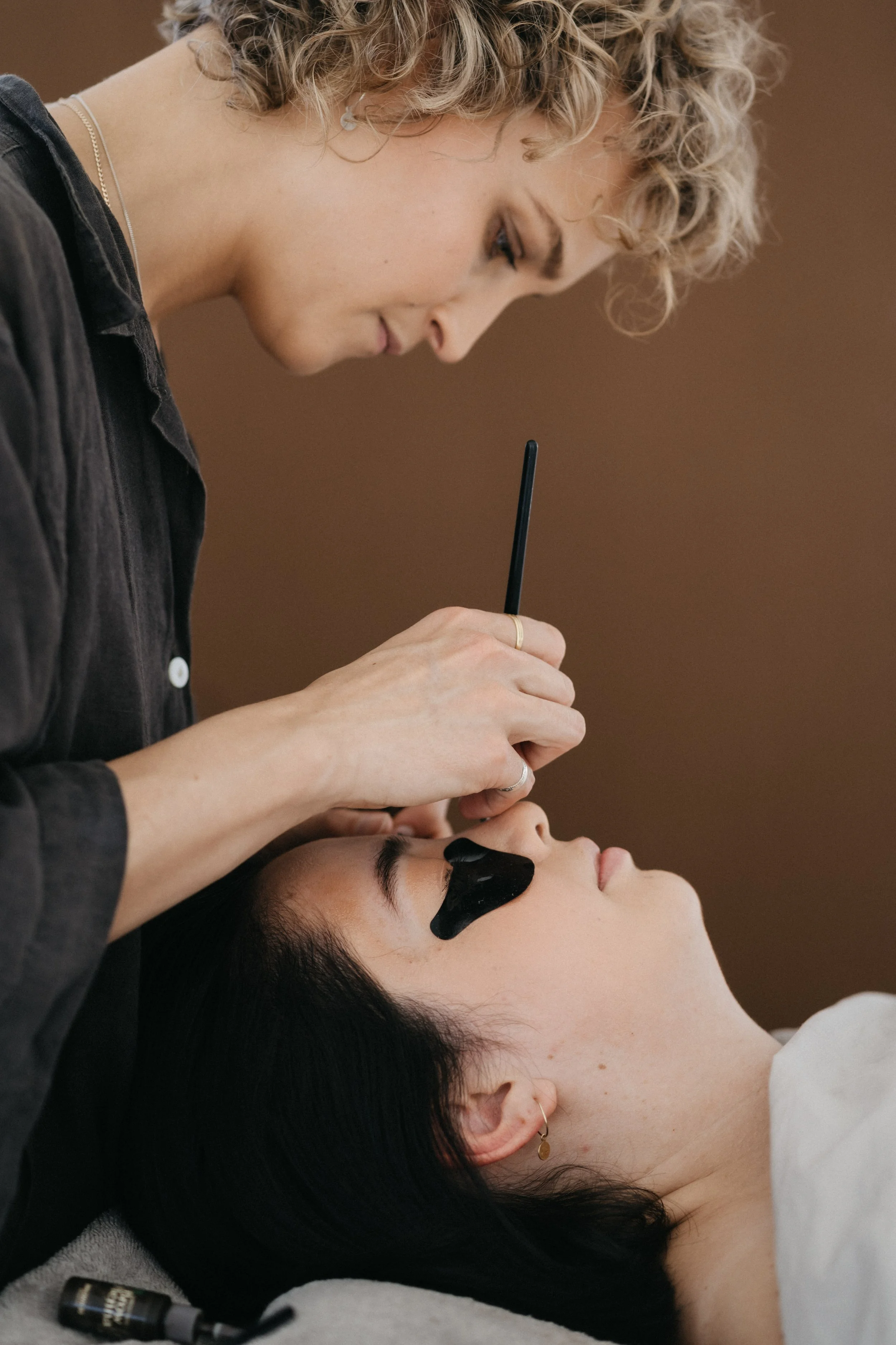A person with black hair lying down with black face paint around their eyes, receiving makeup or special effects application from a woman with curly blonde hair, wearing a black jacket and jewelry, in a makeup or special effects studio.