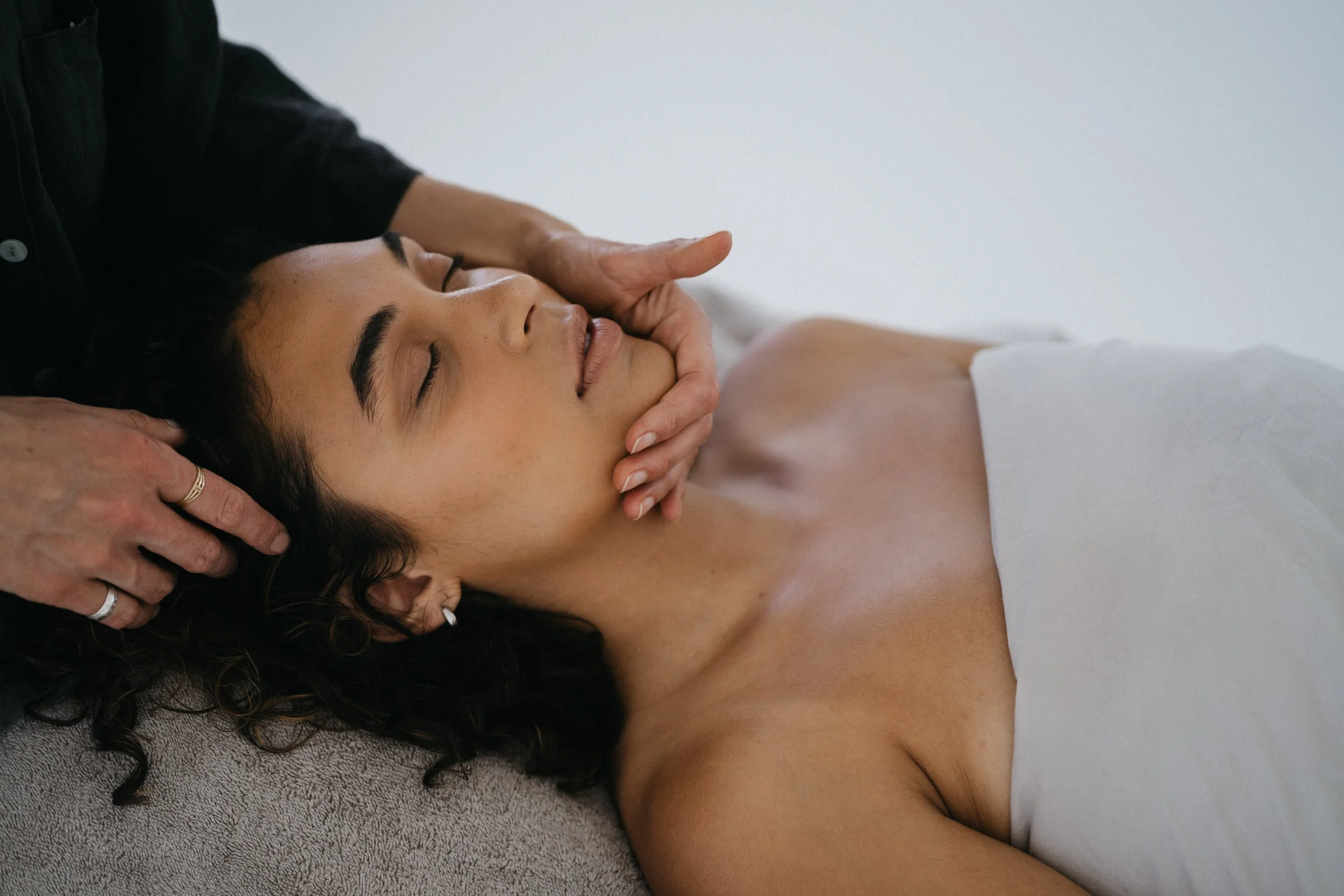 A woman lying down with her eyes closed receiving a massage on her neck and shoulders from a massage therapist.