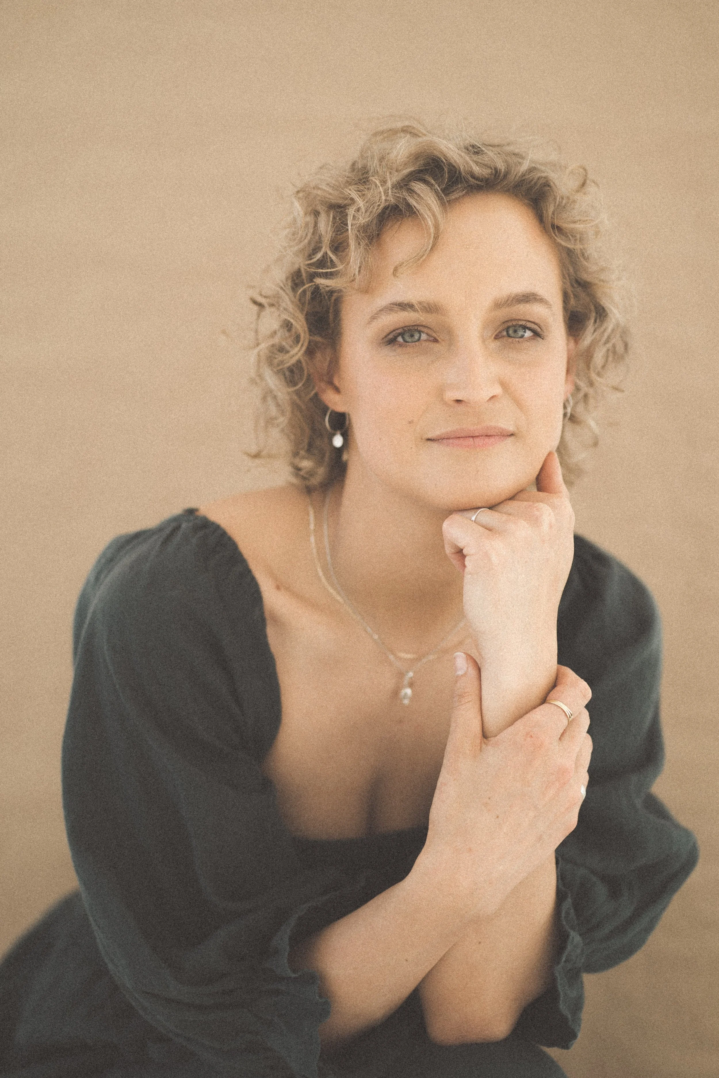 A woman with short curly blonde hair and blue eyes, wearing a black top with puffed sleeves. She has jewelry including earrings, a necklace, and rings, and is resting her chin on her hand with a gentle smile, against a beige background.
