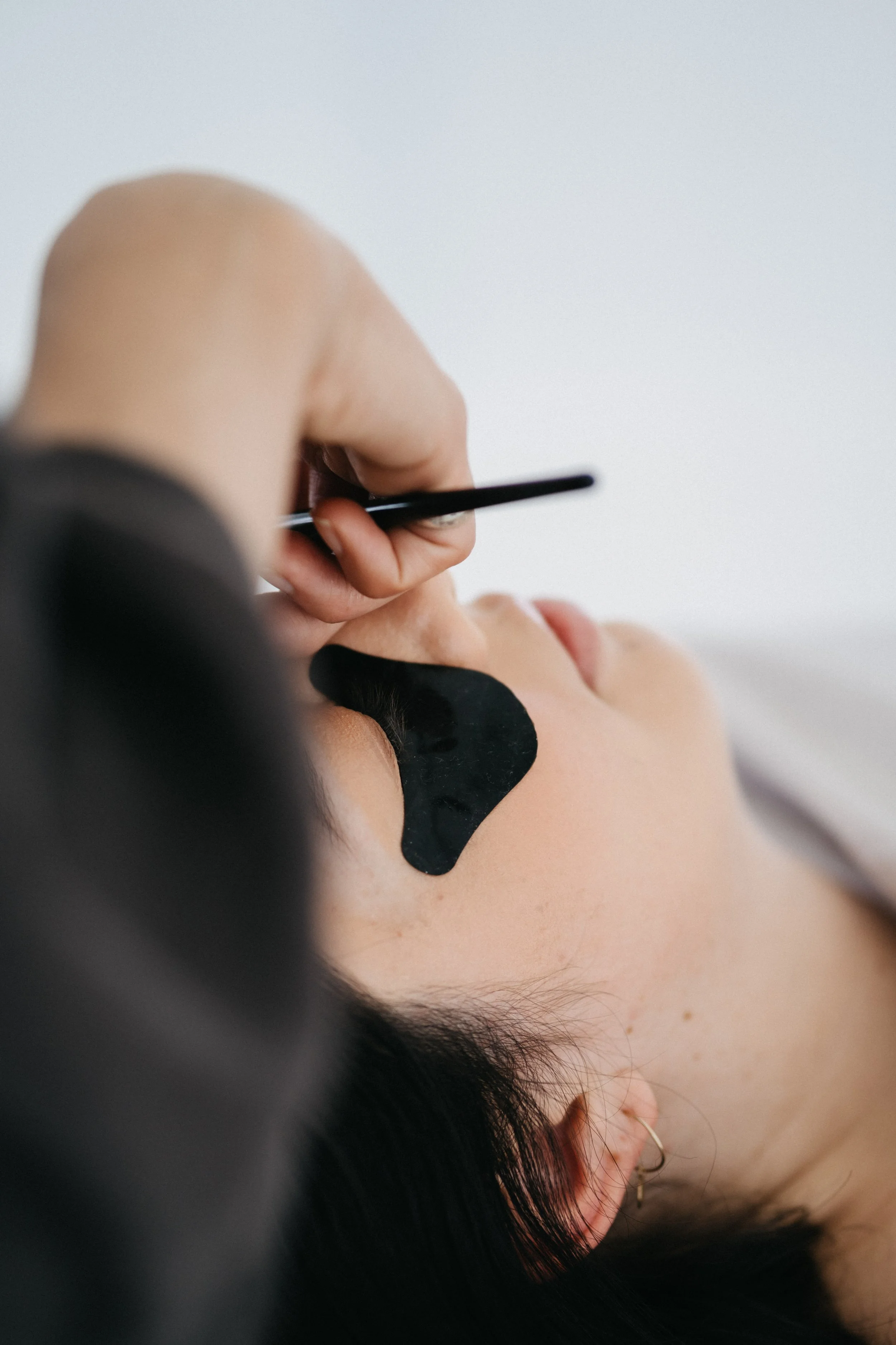 Person receiving a facial with a black facial mask applied, lying down with eyes closed, and another person holding a tool near their face.