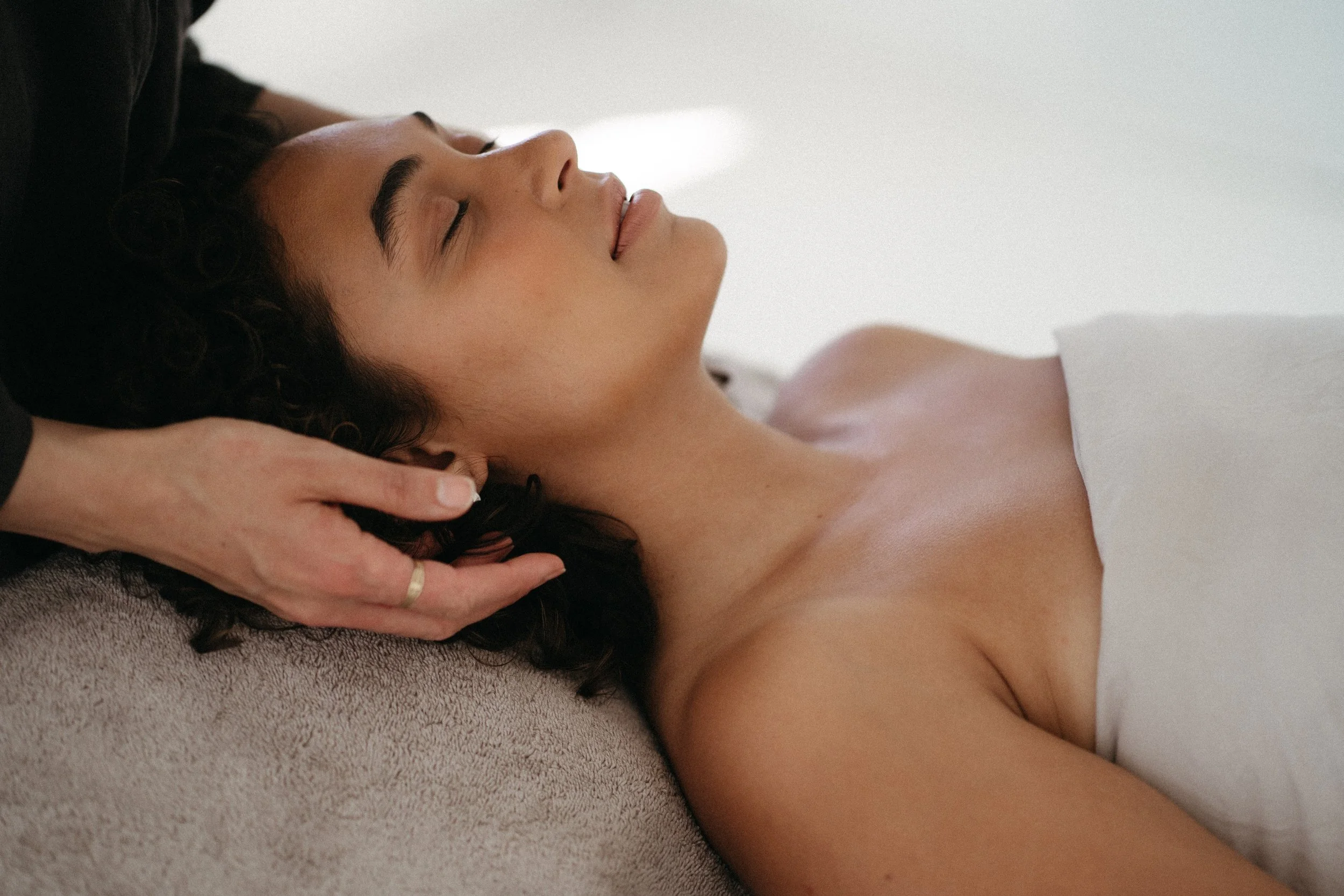 A woman with curly dark hair receiving a massage, lying on her back on a massage table with her eyes closed.