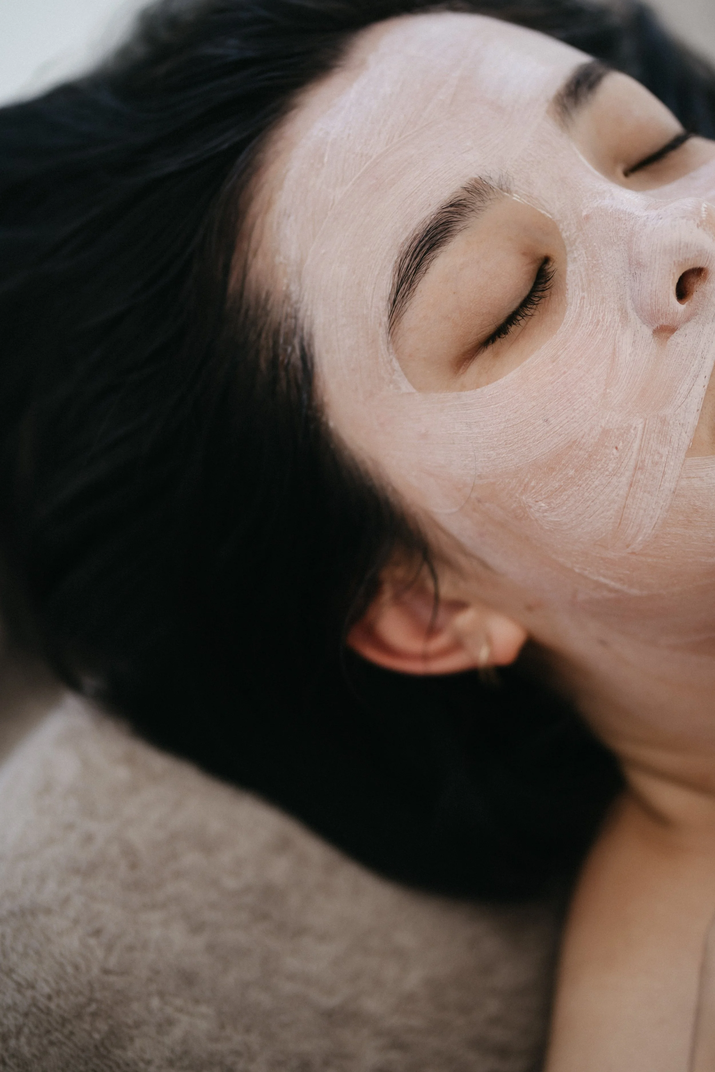 Close-up of a woman with dark hair and light skin, lying down with her eyes closed, wearing a facial mask.