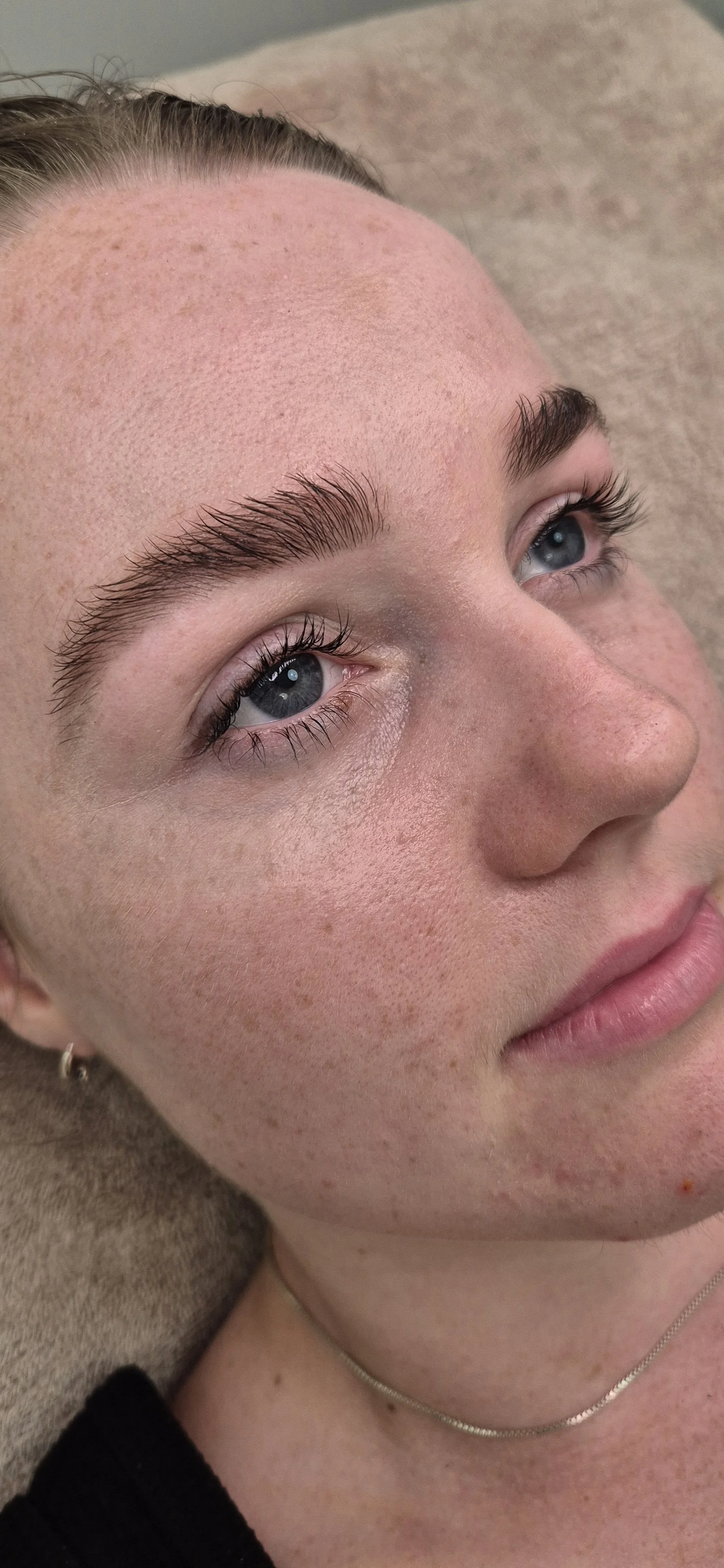 Close-up of a woman's face with makeup, blue eyes, and a silver necklace, lying on a beige surface.