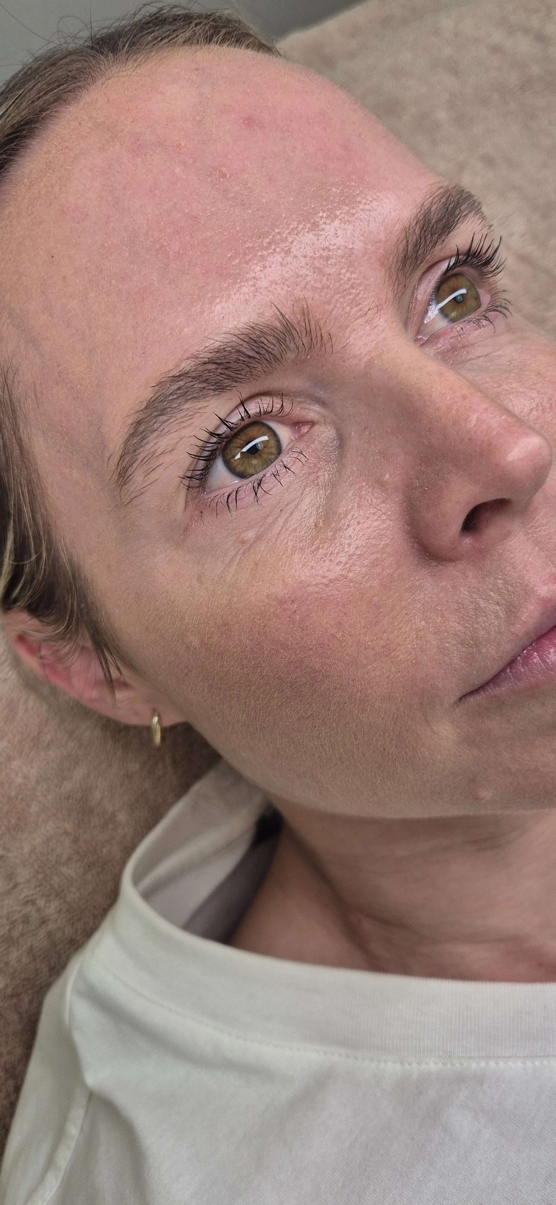 Close-up of a woman's face, showing her with light skin, hazel eyes, arched eyebrows, and a small gold hoop earring, while lying on a soft surface.