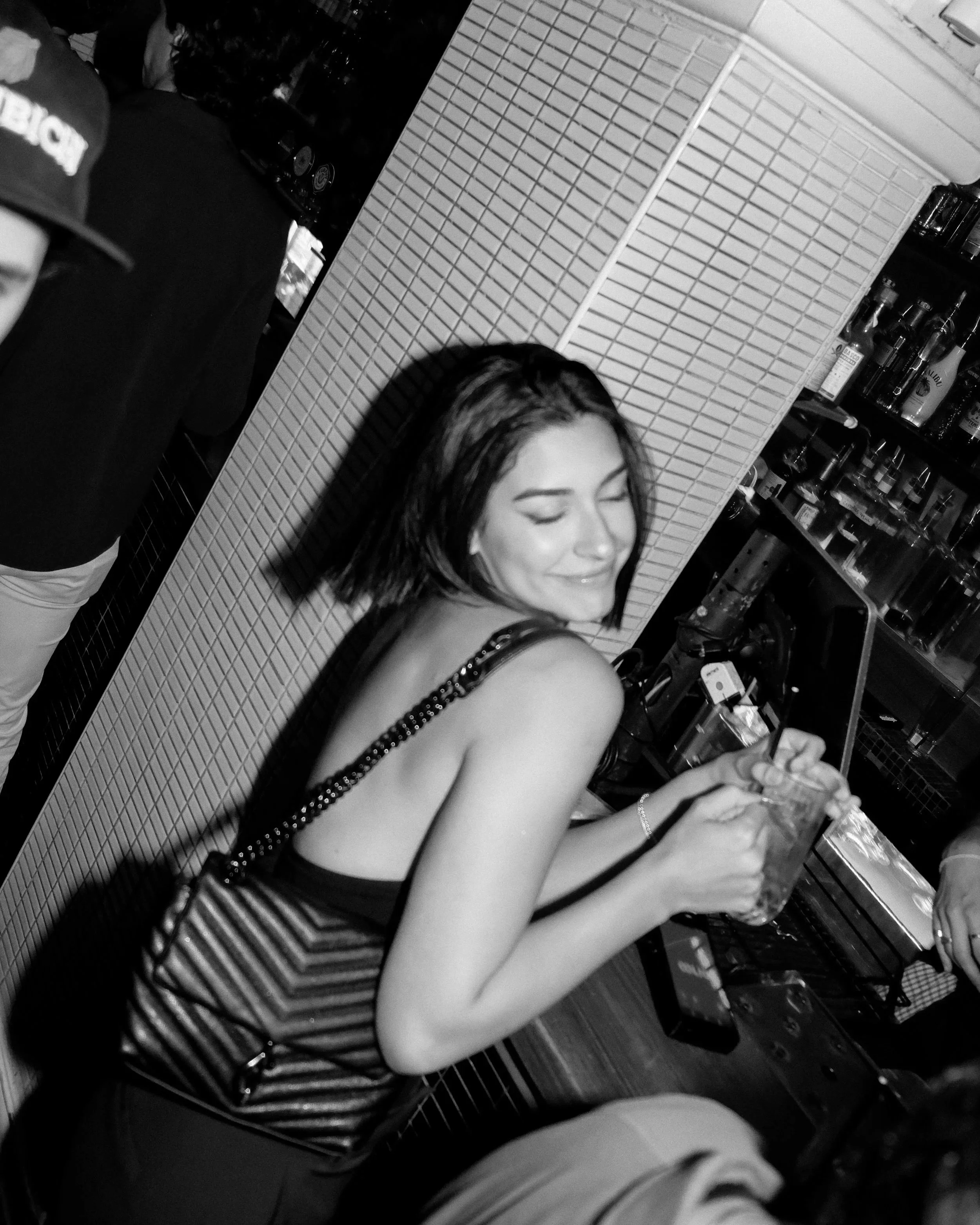A young woman with long dark hair and a beaming smile sitting at a bar or counter. She is wearing a sleeveless top with chain straps and appears to be holding a drink with a straw. In the background, there are shelves filled with bottles.