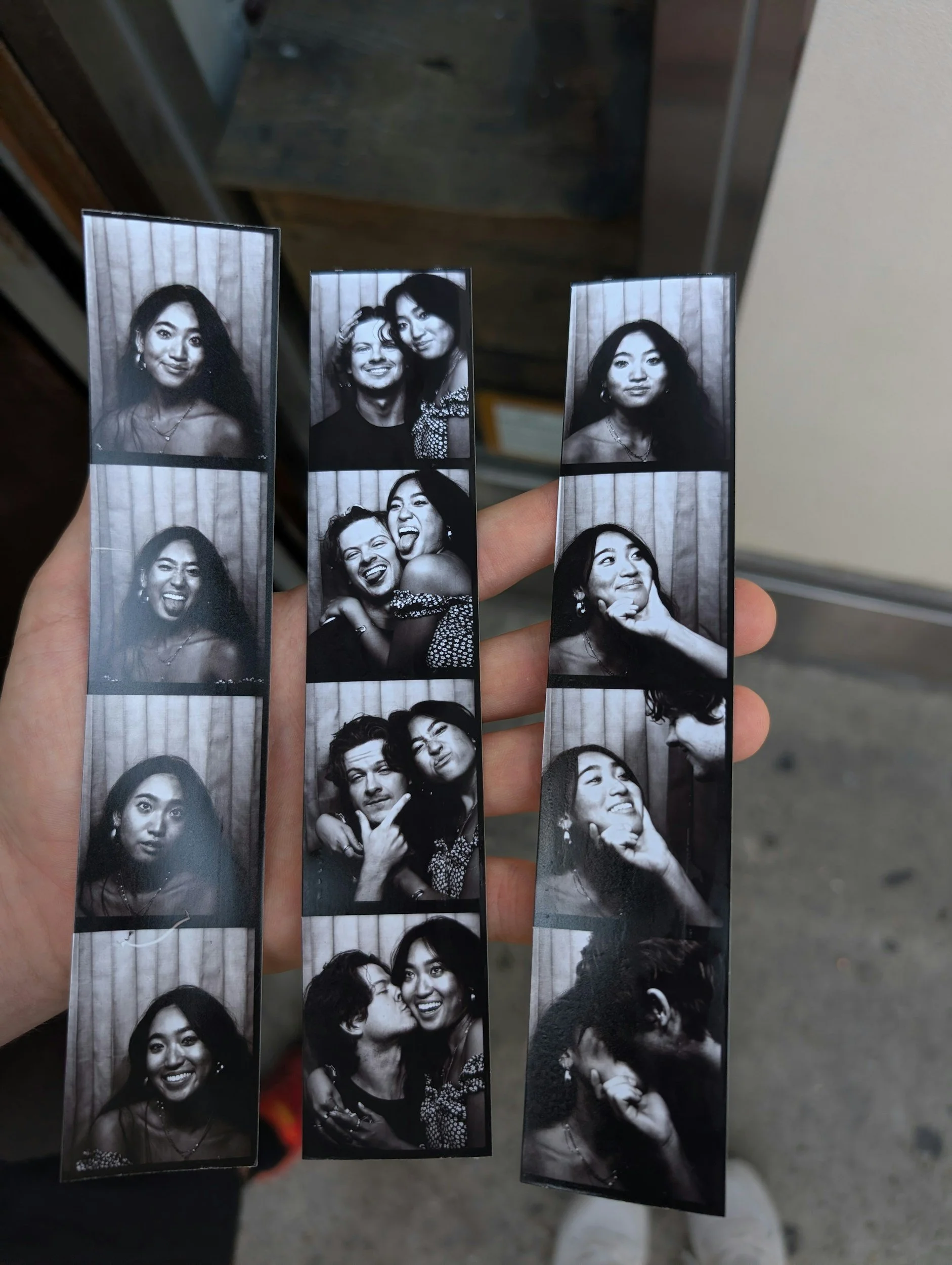 A hand holding three strips of photo booth pictures in black and white, showing multiple images of two women and a man, with a background of vertical striped curtains.