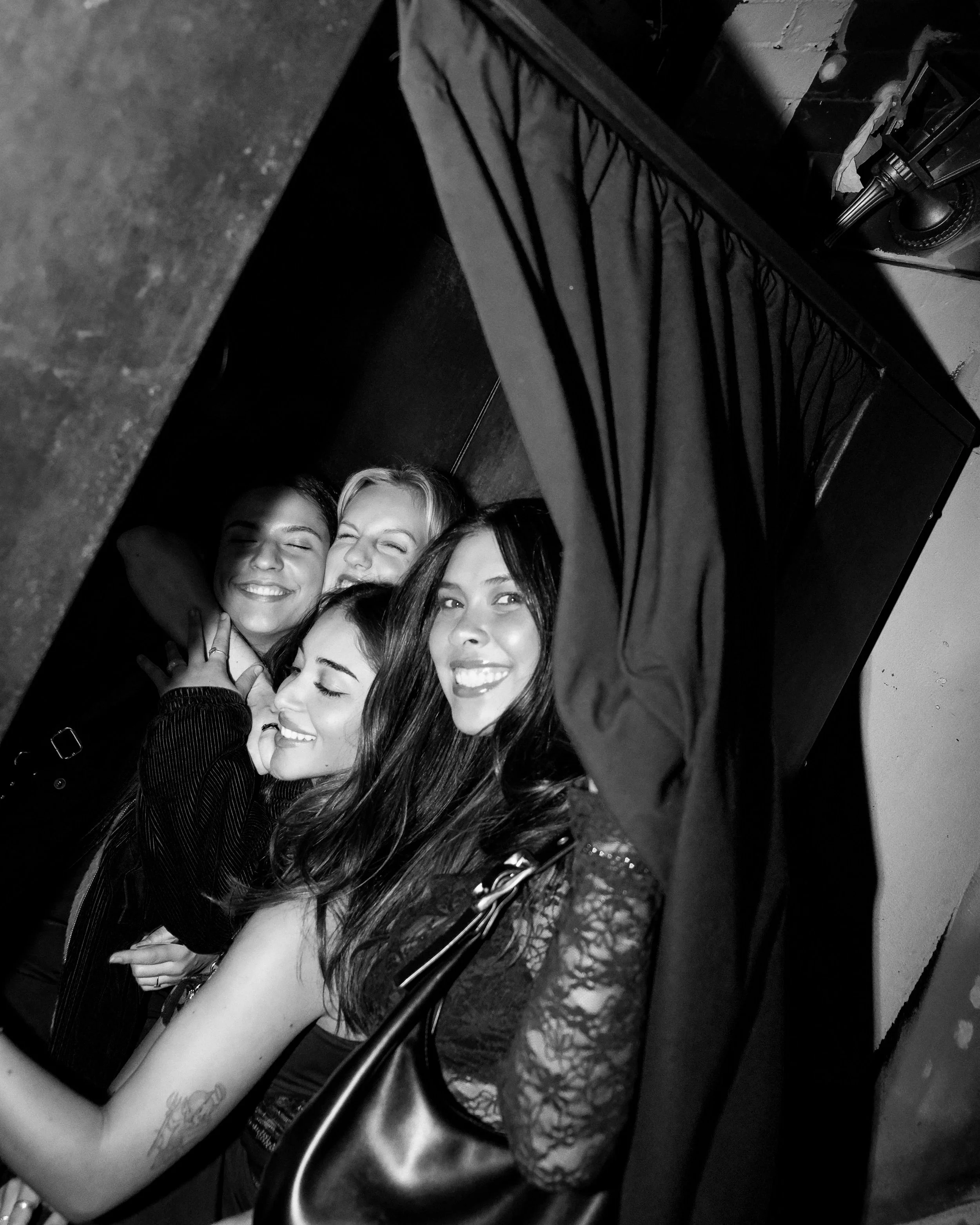 Black and white photo of five happy women with arms around each other, smiling, closely gathered, lying on the floor with a dark background inside a photobooth in melbourne.