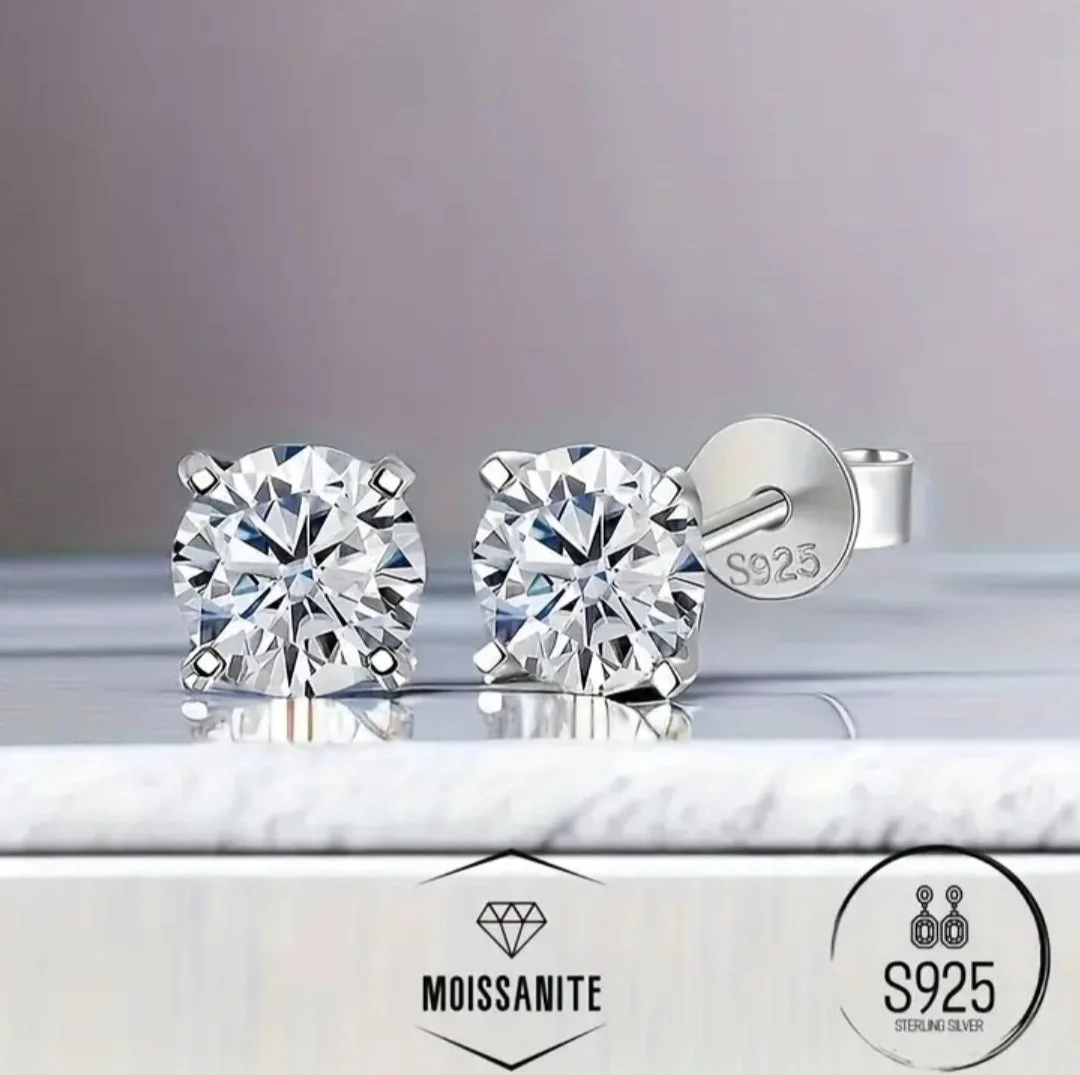 Pair of silver stud earrings with large round clear gemstones, stamped S925, and displayed on a marble surface with the brands Moissanite and S925 Sterling Silver.