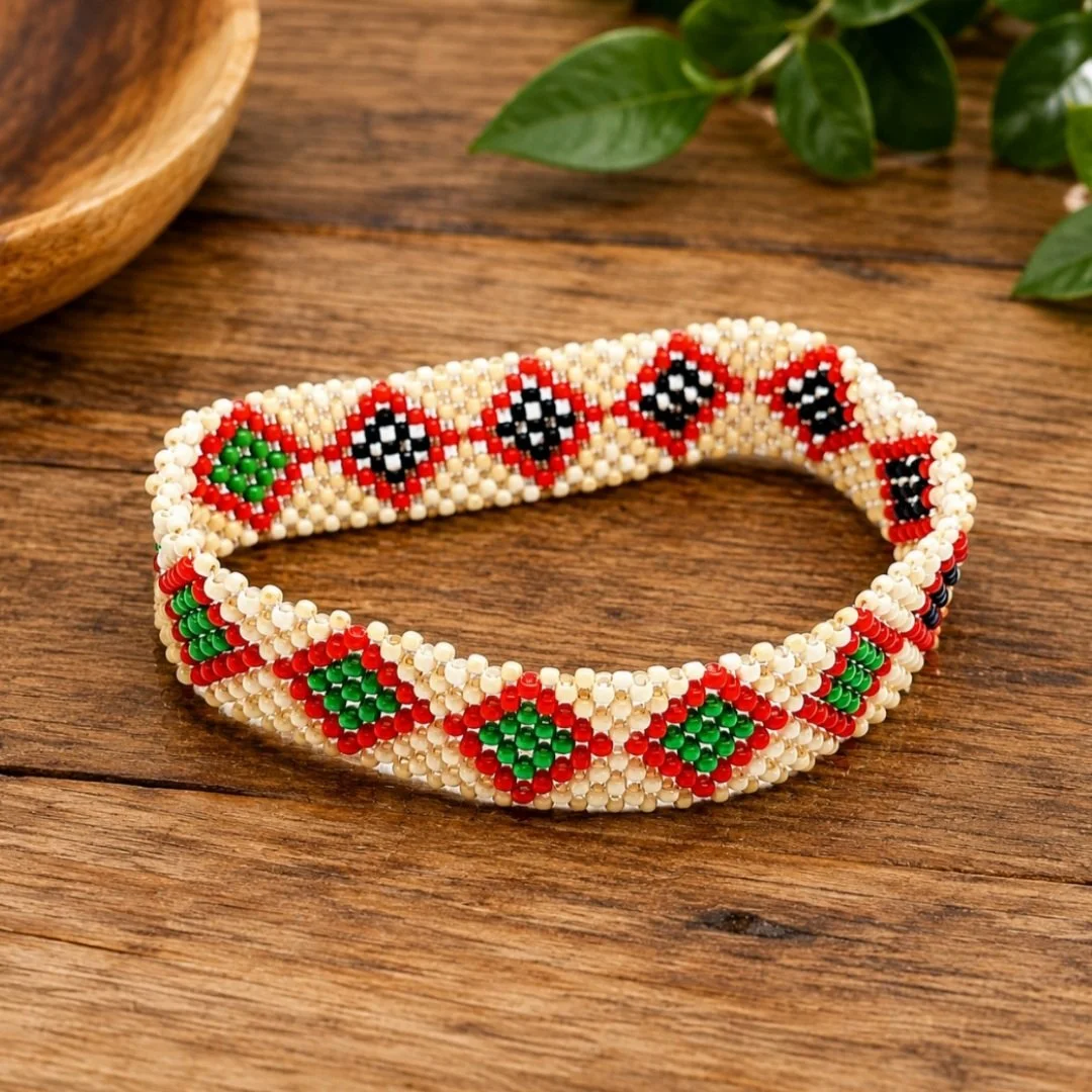 Beaded bracelet with red, green, black, and cream diamond patterns on a wooden surface with green leaves in the background.