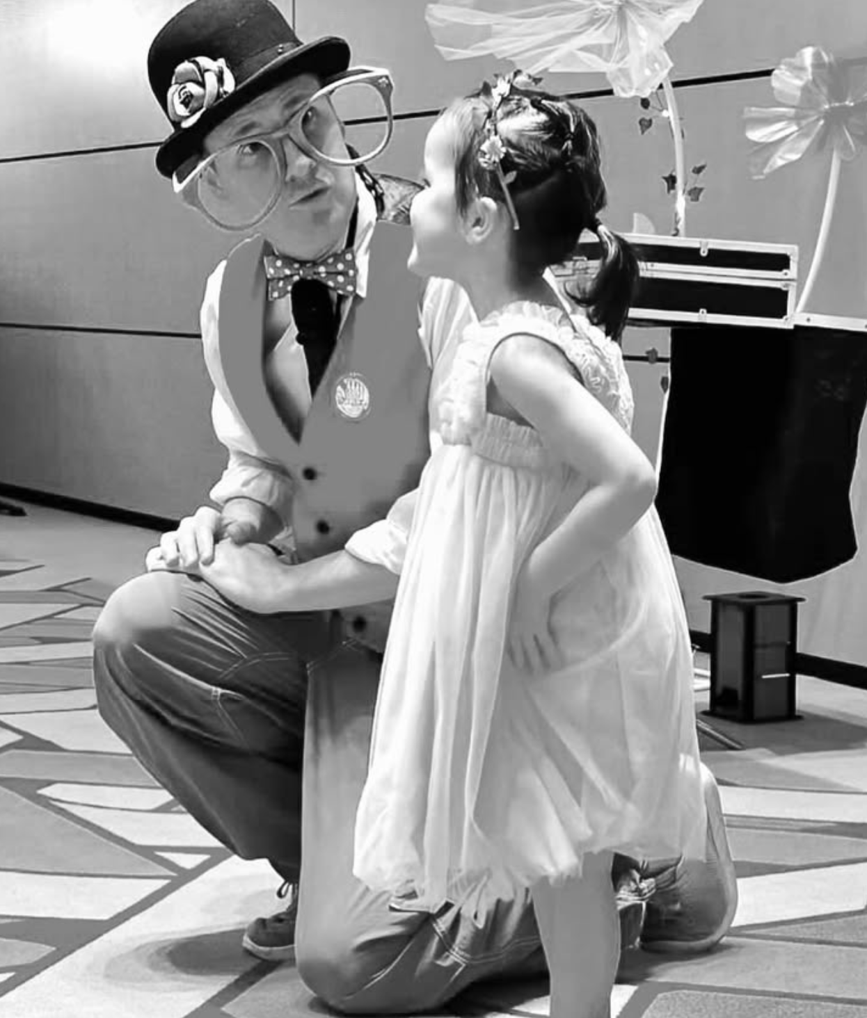 dressed as a clown in oversized glasses, a hat with a flower, and a vest holding hands with a young girl in a white dress with lace sleeves, who is kneeling on a patterned floor. The background shows a wall with panels and some decorations, including paper flowers.